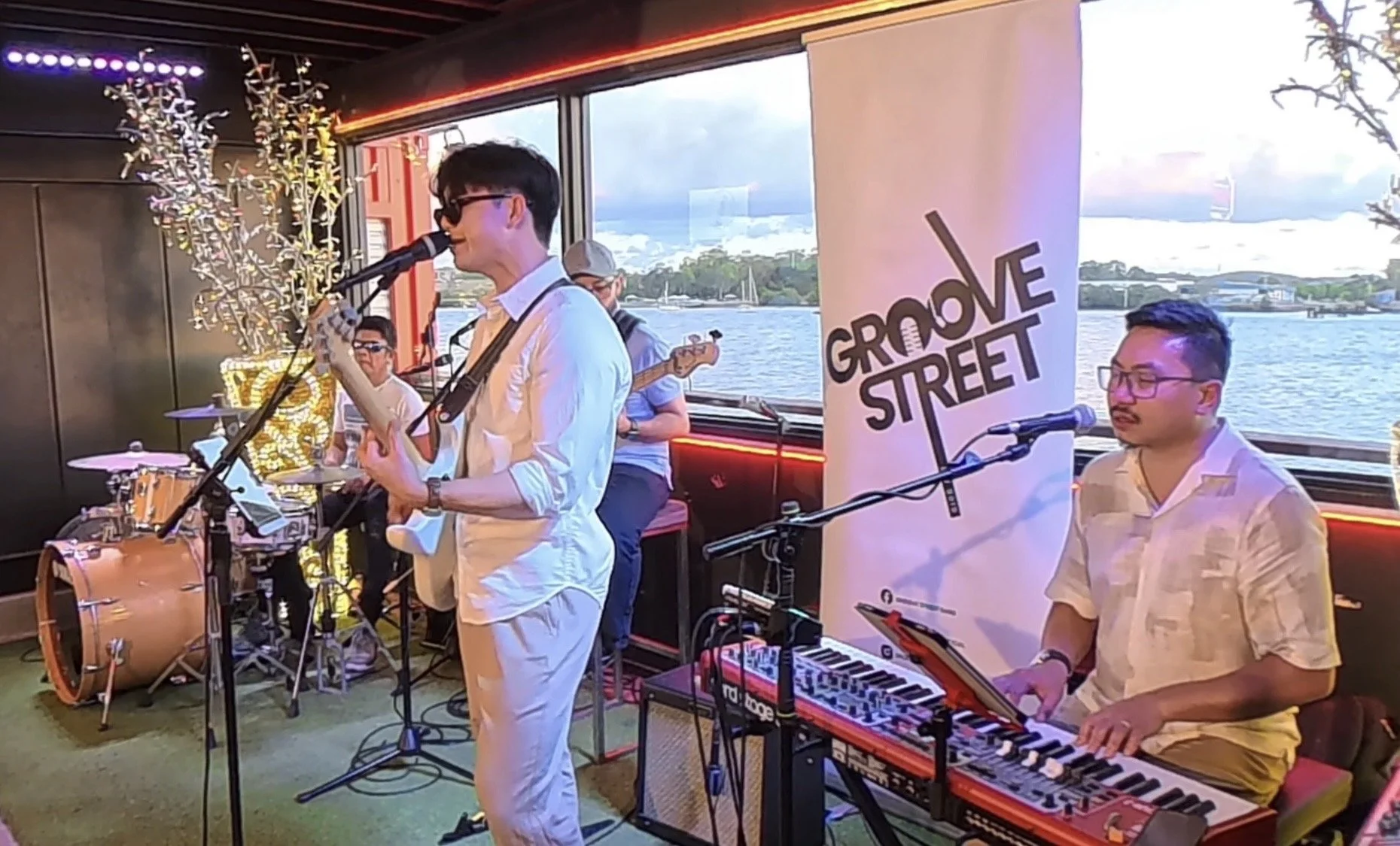 A band performing live on a stage with musical instruments, a keyboard, drums, and microphones, at an event called Groove Street, by a body of water with a scenic view outside the window.