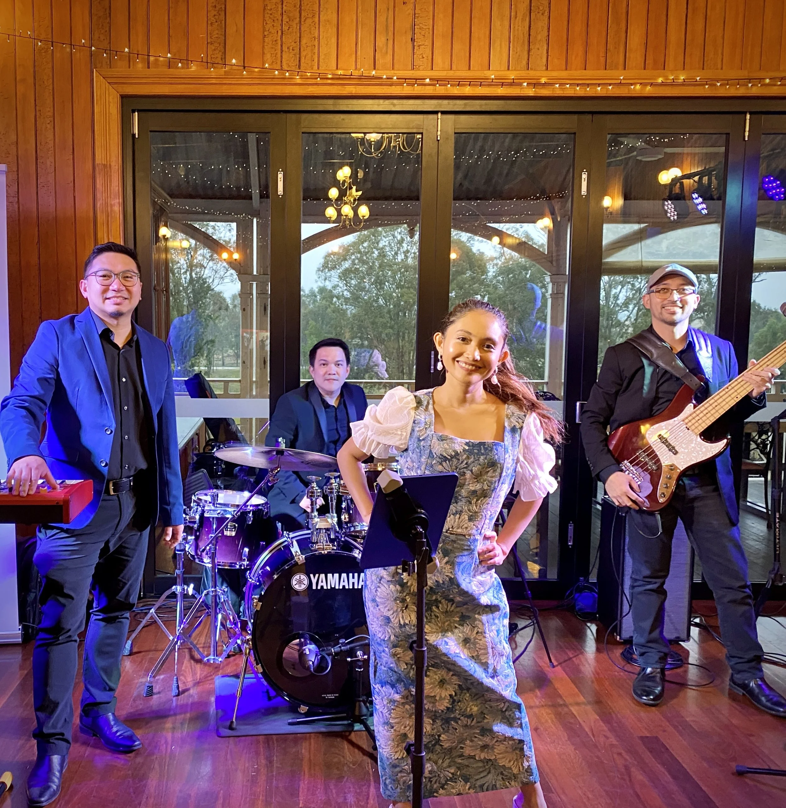 A four-member band performing indoors with large windows and greenery outside. The band includes a keyboard player, a drummer, a female singer, and a bass guitarist. The female singer is smiling and standing at a microphone, wearing a floral dress wi