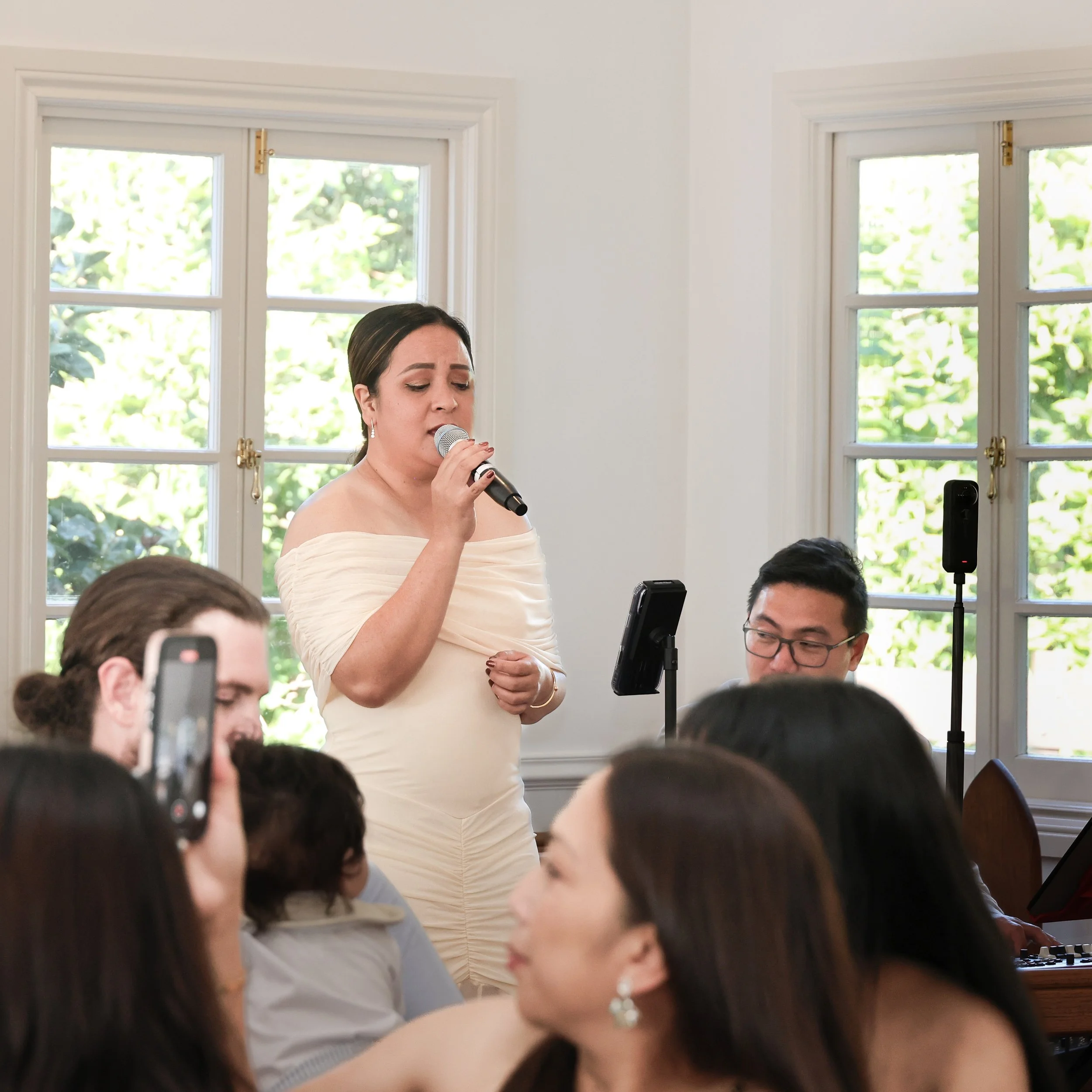 A woman in a white off-the-shoulder dress singing into a microphone during a live performance, with people and a musician in the background in a bright room with large windows.