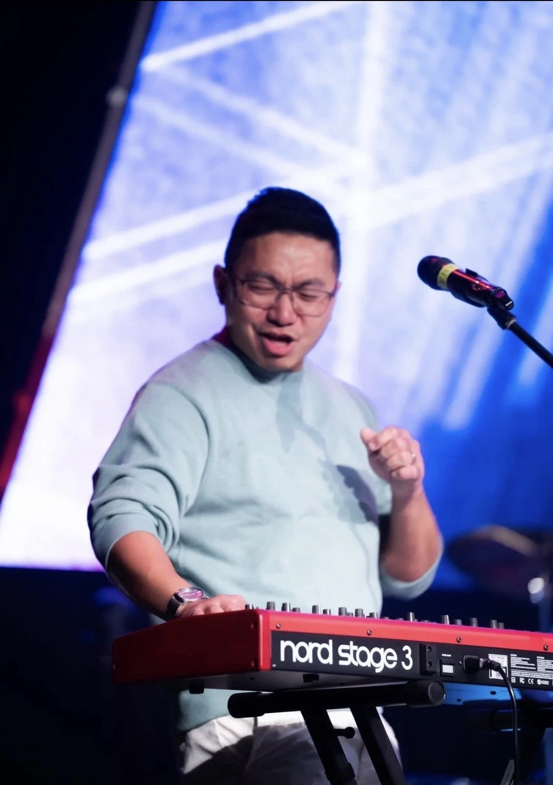 A man with glasses wearing a light-colored sweatshirt, playing a red Nord Stage 3 keyboard on stage, with a microphone nearby, and a blue-lit background.