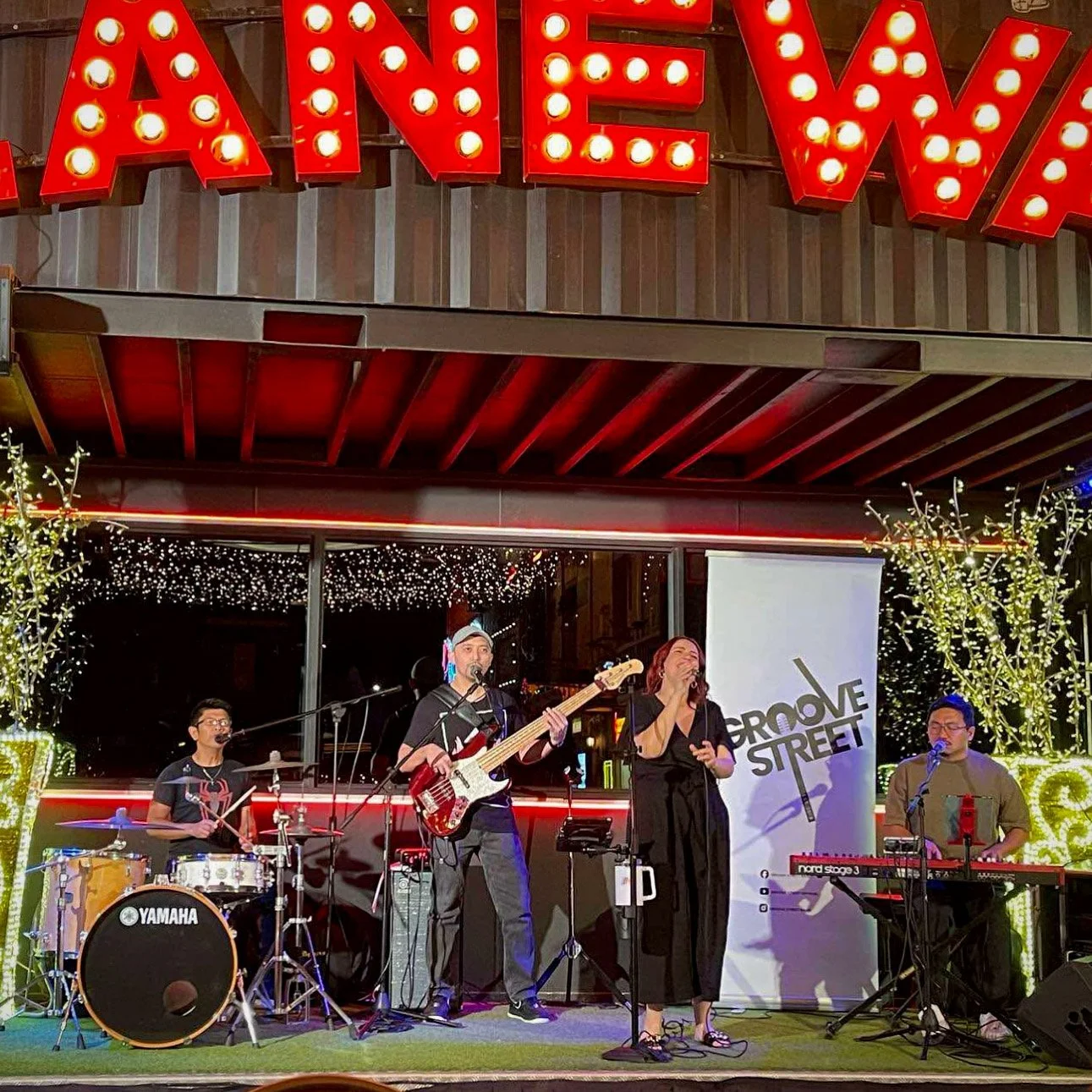 A band performing live on stage in front of a sign that says "Amazing" with illuminated letters. The band consists of a drummer, a bassist, a female vocalist, and a keyboardist. The stage is decorated with string lights and trees with white string li