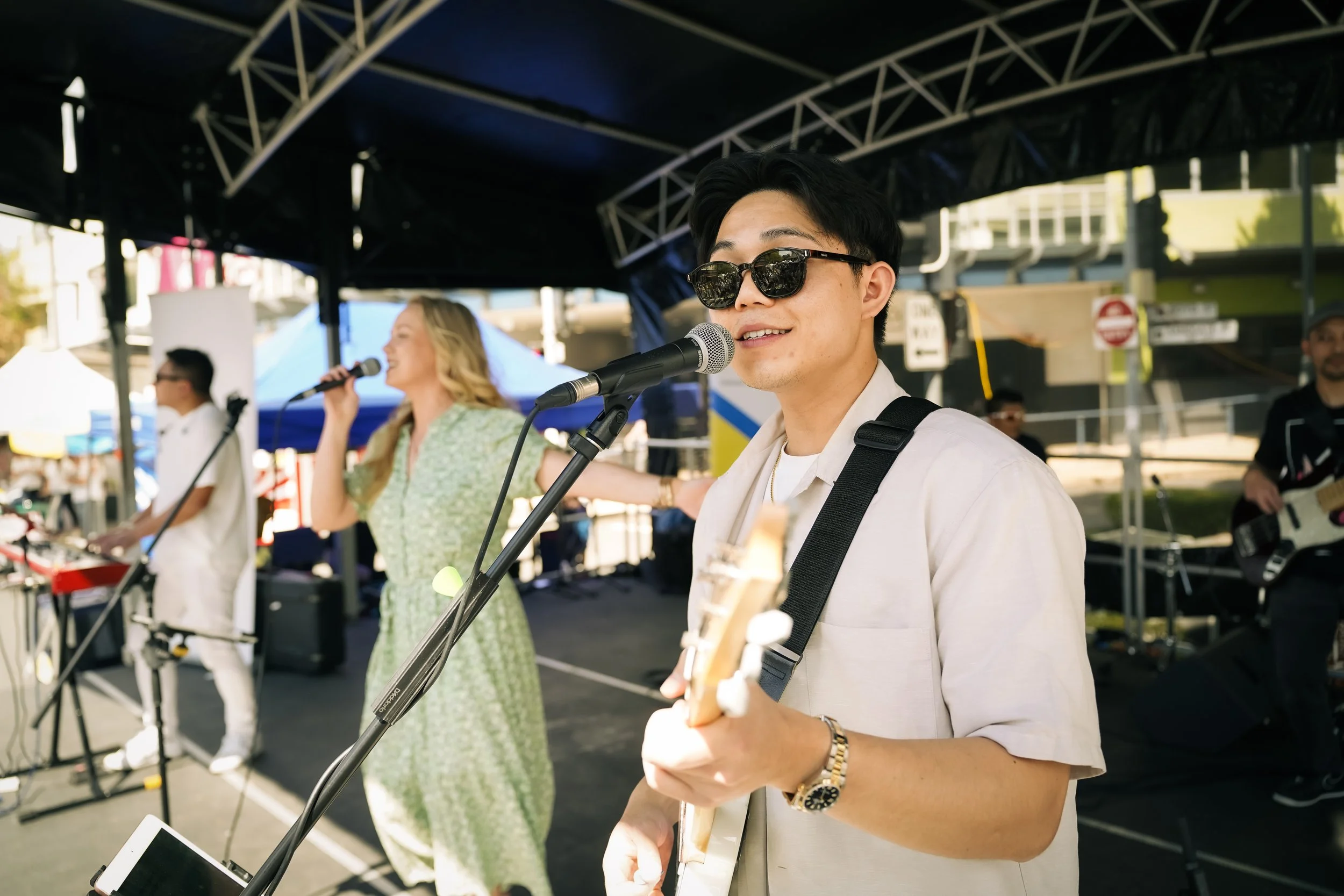 Musicians performing on an outdoor stage, with a young man playing guitar and wearing sunglasses, a woman singing into a microphone, and other band members playing instruments, under a canopy.