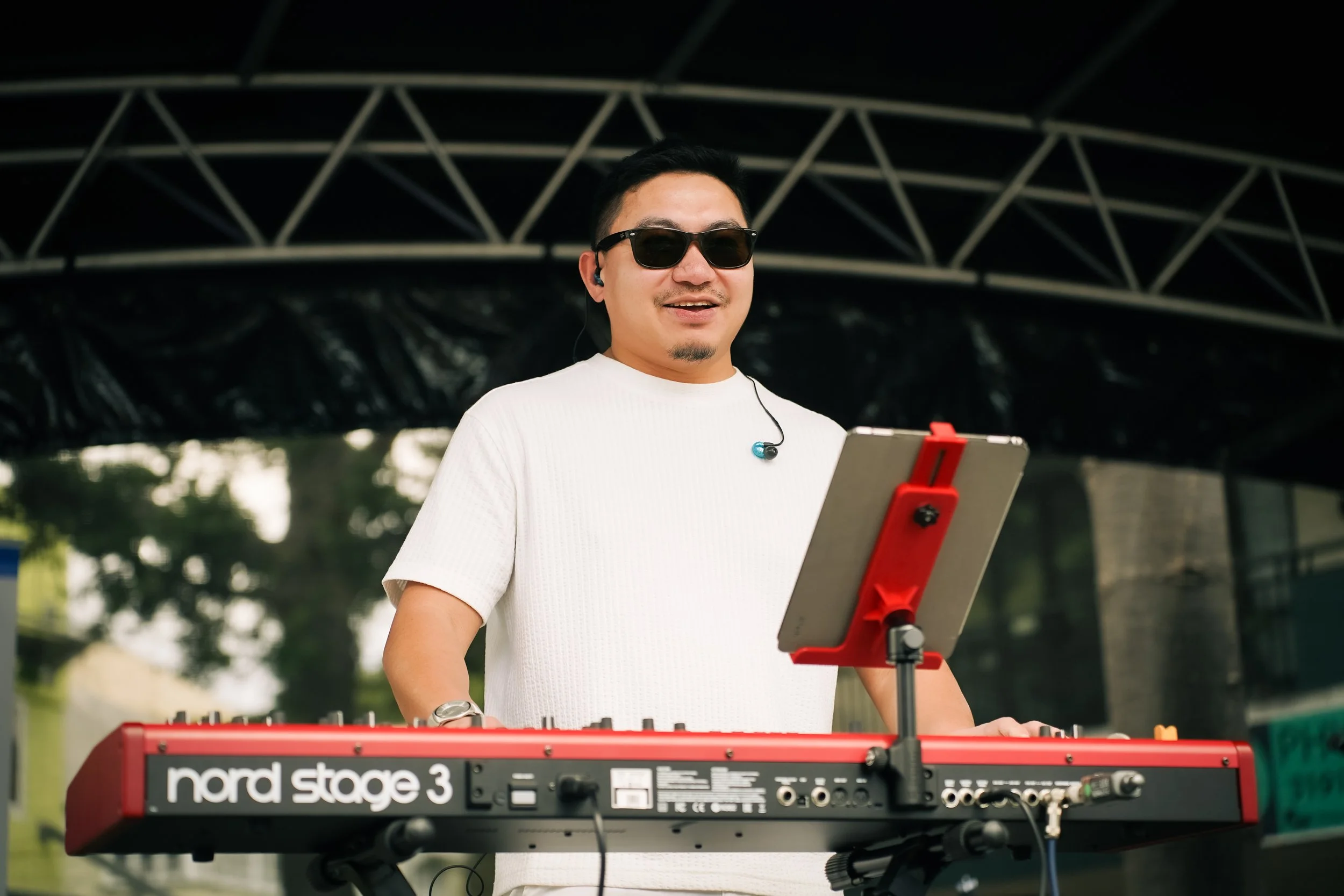 Man wearing sunglasses and headphones playing a red Nord Stage 3 keyboard outdoors.