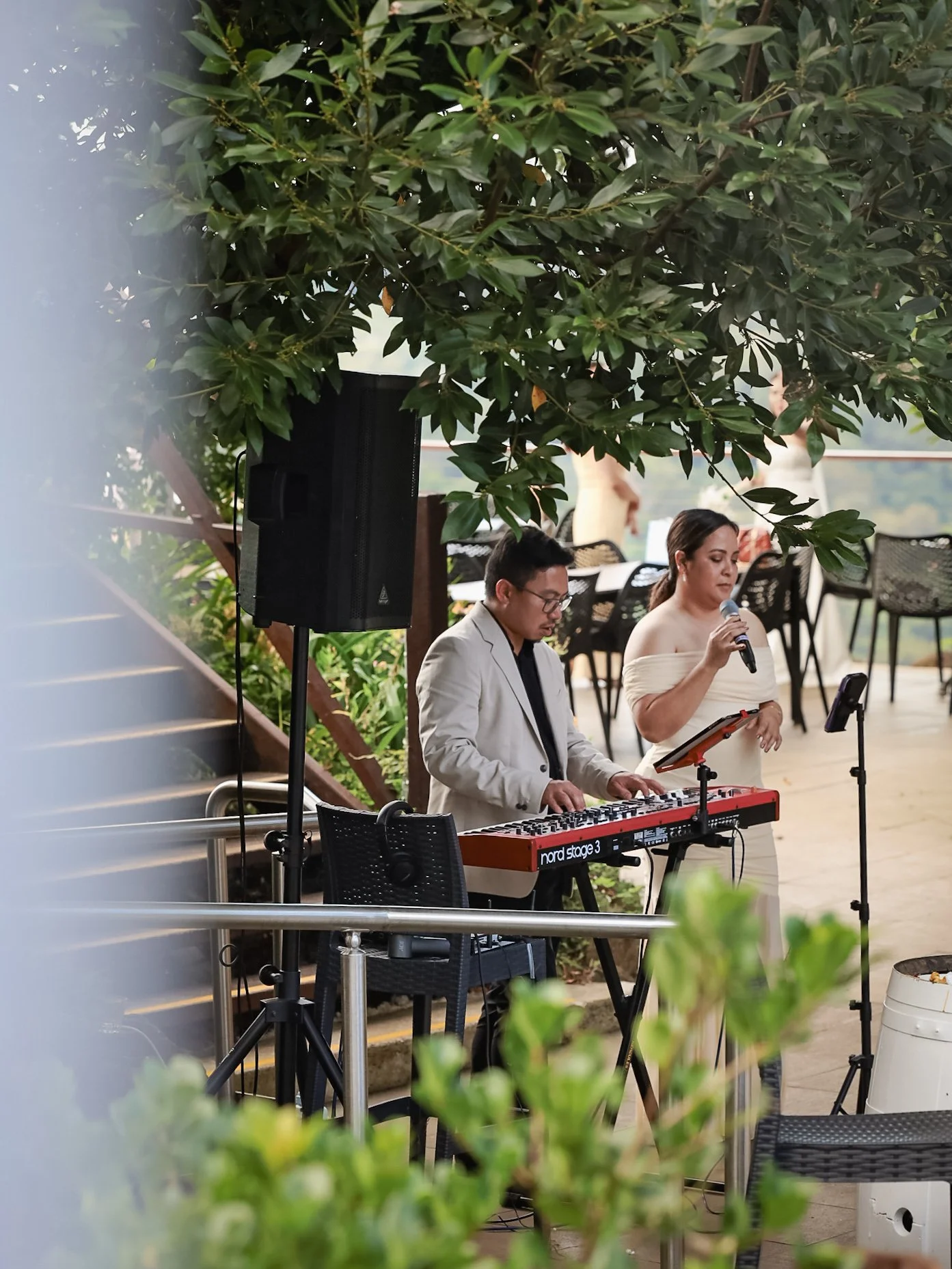 A musician playing a keyboard and a singer with a microphone performing outdoors on a stage surrounded by greenery.