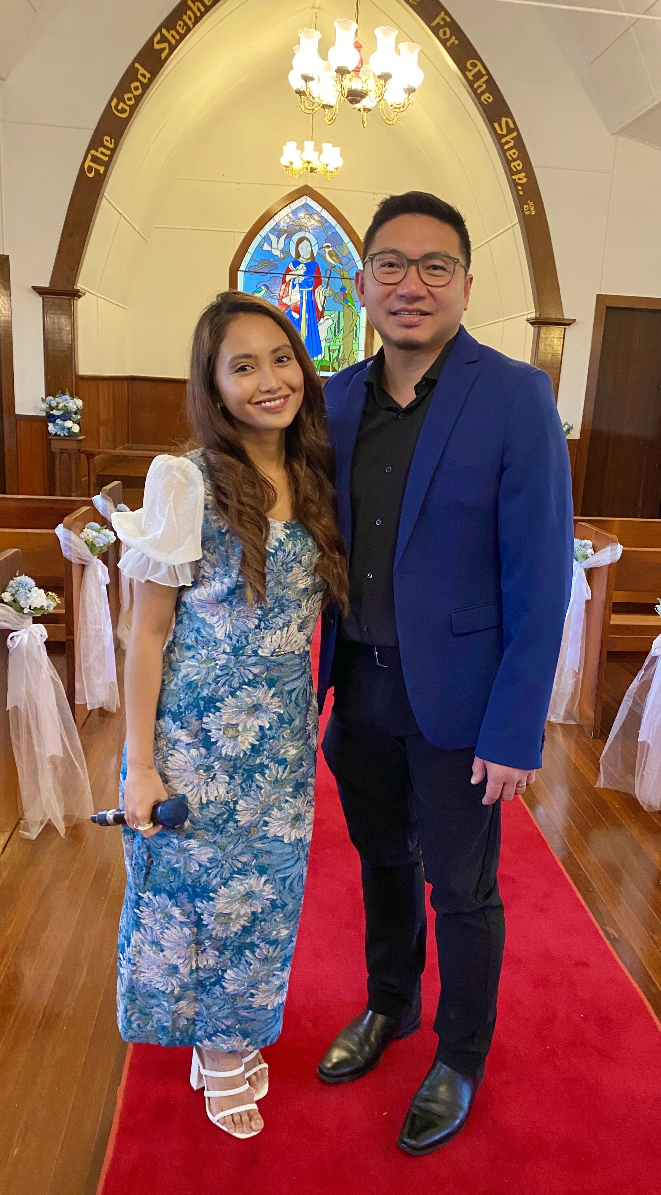 A man and woman standing inside a church with wooden pews decorated with flowers and ribbons, a colorful stained glass window with a religious figure of Jesus Christ behind them, and a chandelier hanging from the ceiling. The woman is holding a micro