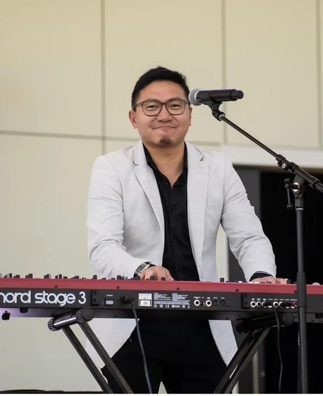 A man smiling and playing a red and black keyboard with a microphone on a stand in front of him.
