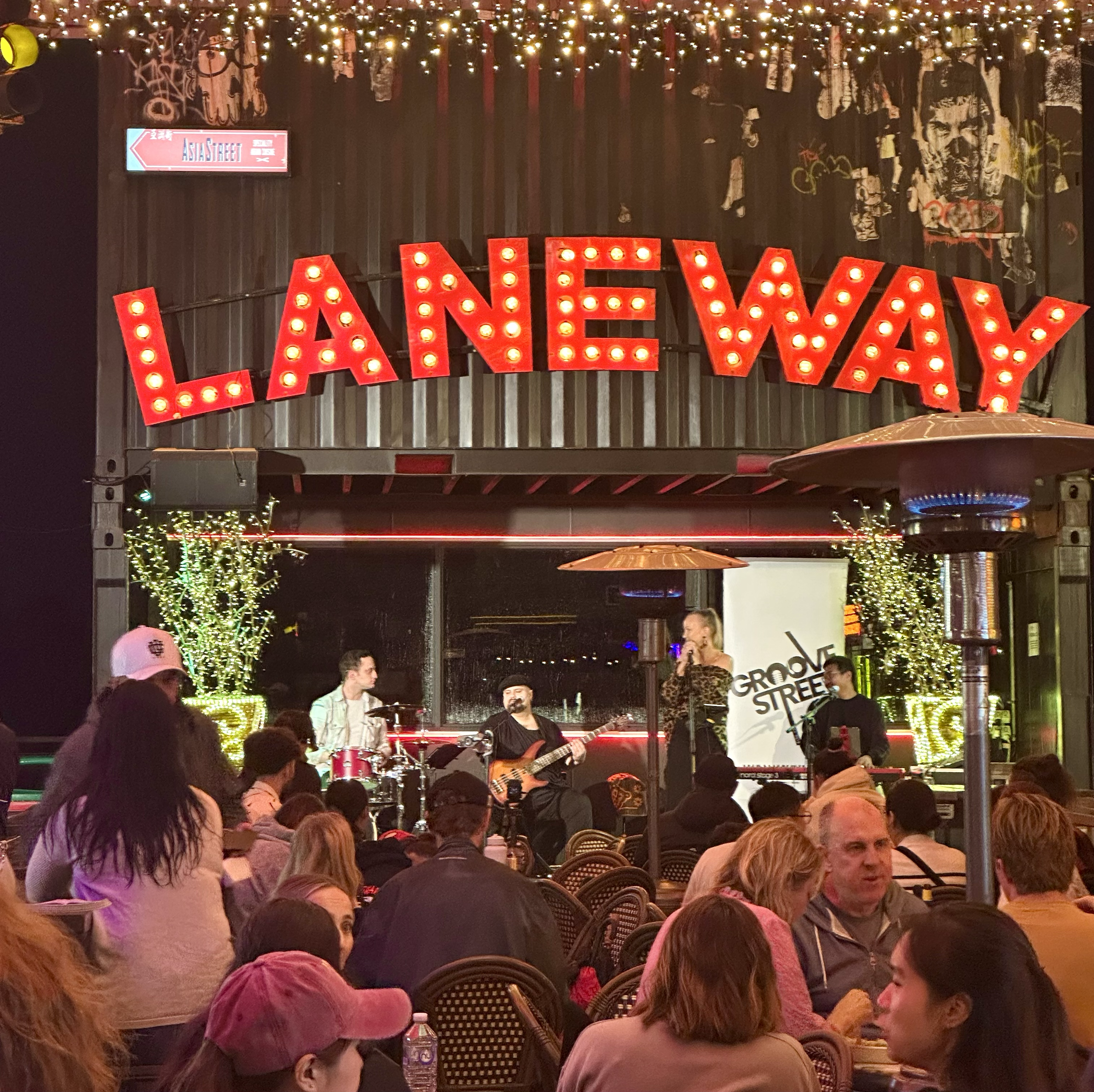 Live music performance at LANE WAY with a band playing on stage and an audience seated at tables, decorated with string lights and illuminated trees.
