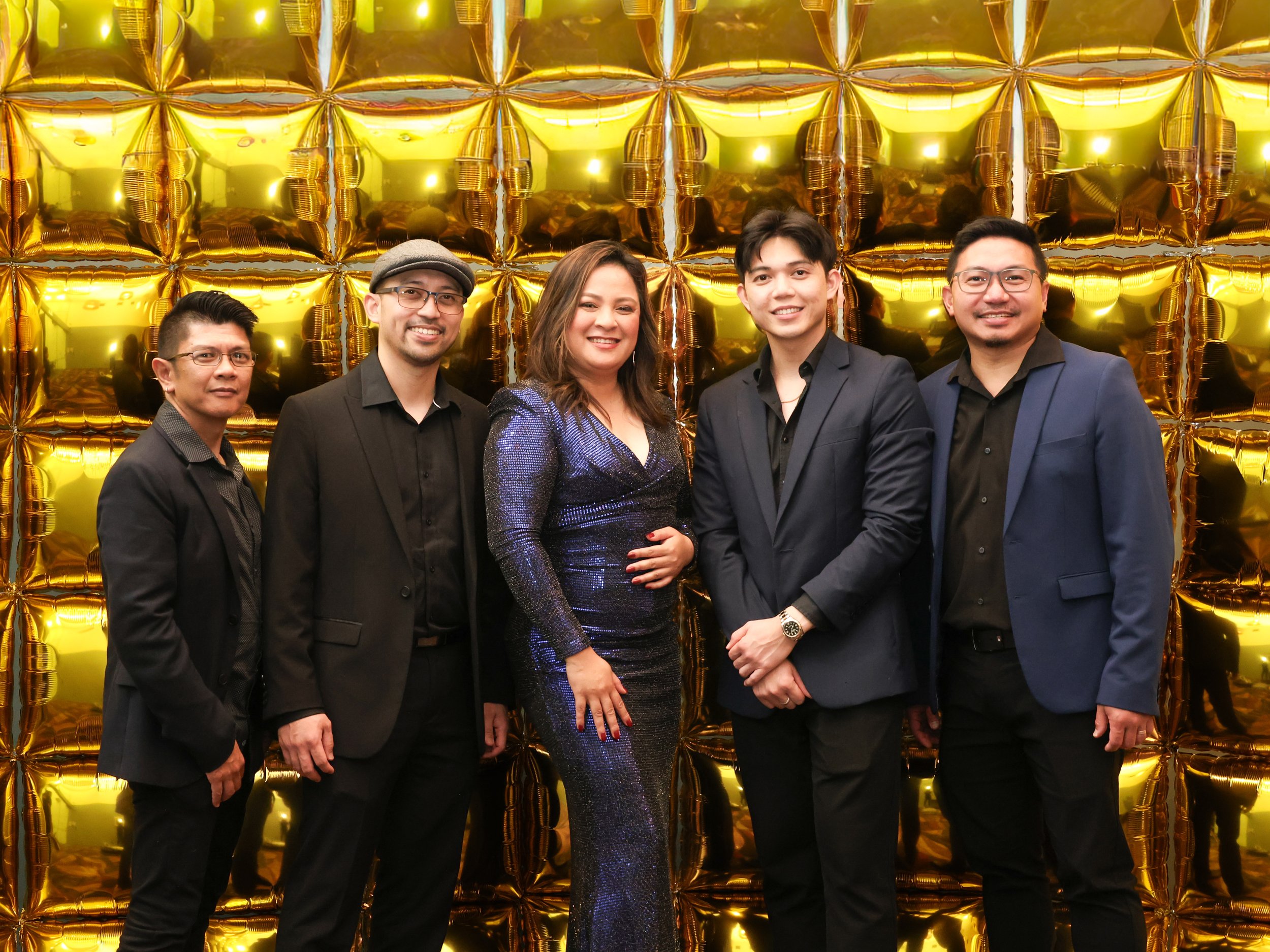 Six people dressed in formal attire, standing in front of a gold-colored balloon backdrop, smiling for the camera at an event or celebration.