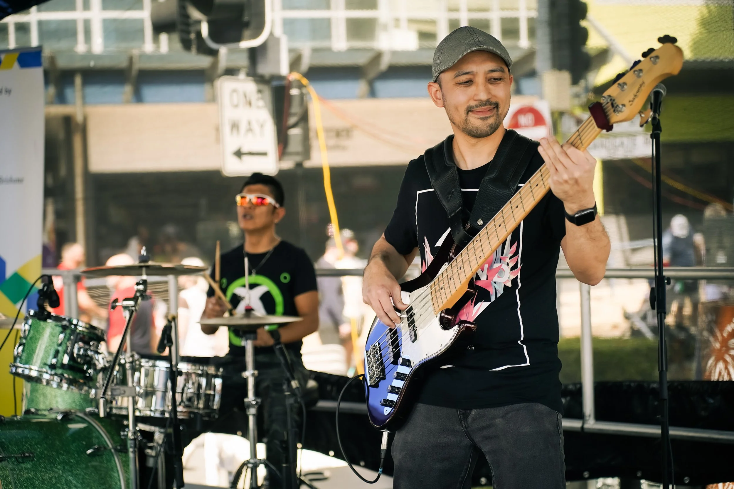 A musician playing electric bass guitar and a drummer playing drums performing outdoors at a music event.