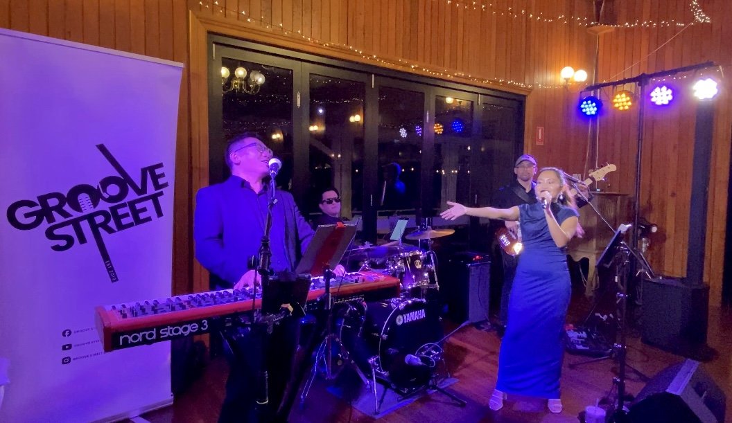 A band named Groove Street performing on stage with a keyboard, drum set, and a singer in a blue dress, in a wooden-paneled venue with colorful lighting.