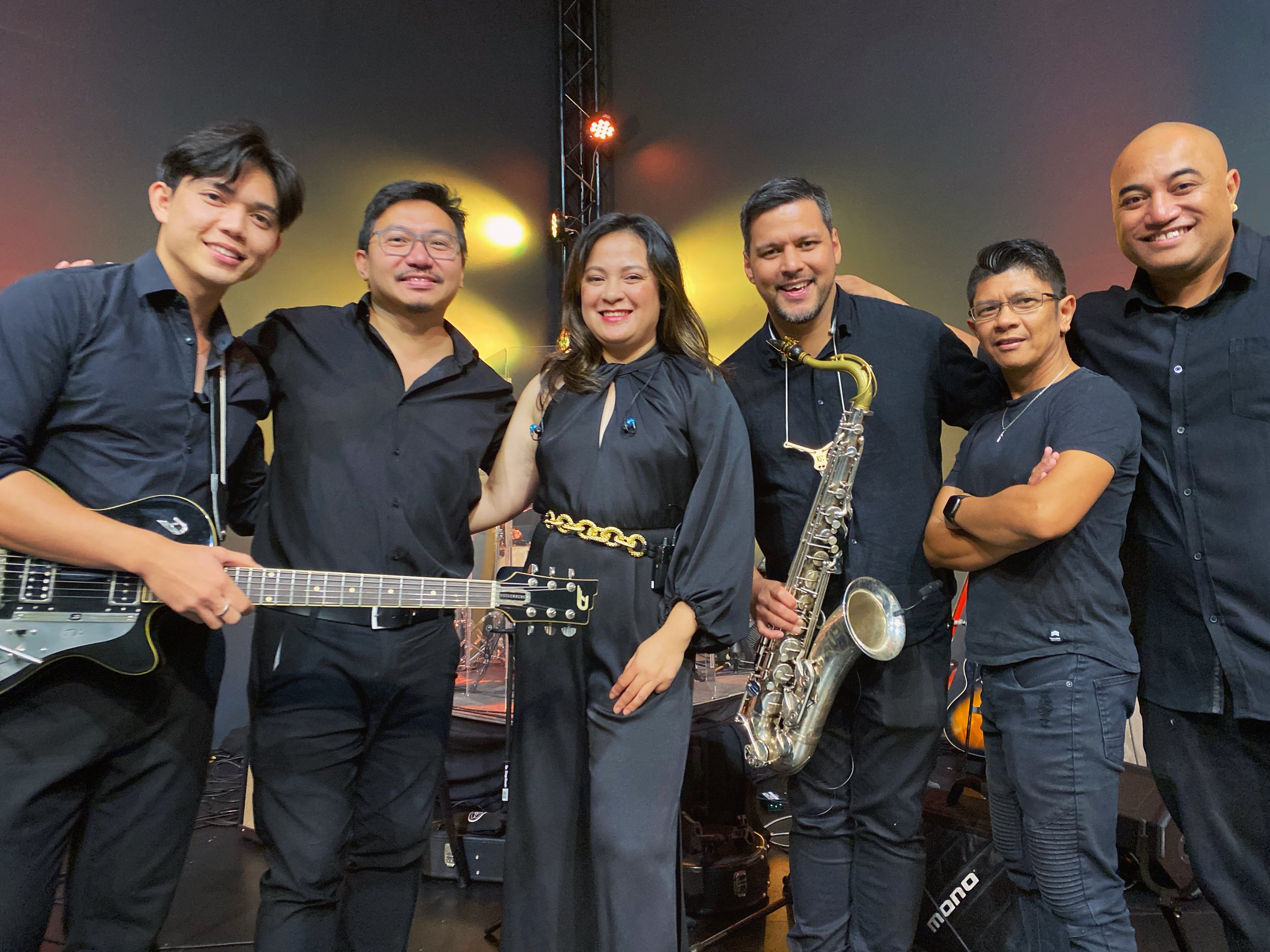 Group of seven musicians on stage, holding their instruments, smiling, with colorful stage lighting in the background.