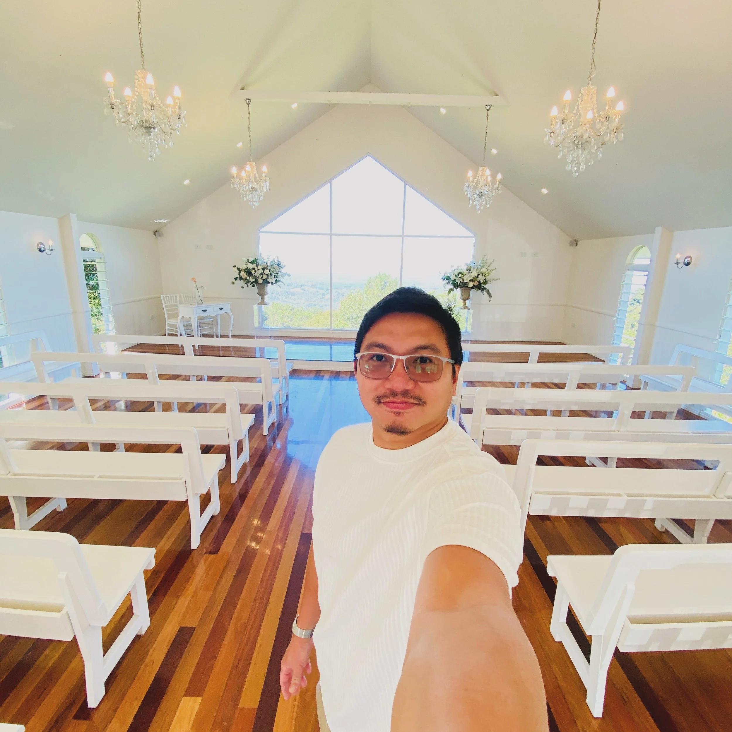 Selfie of a man wearing glasses and a white shirt inside a bright, decorated room with white benches, chandeliers, large window, hardwood floor, and floral arrangements.