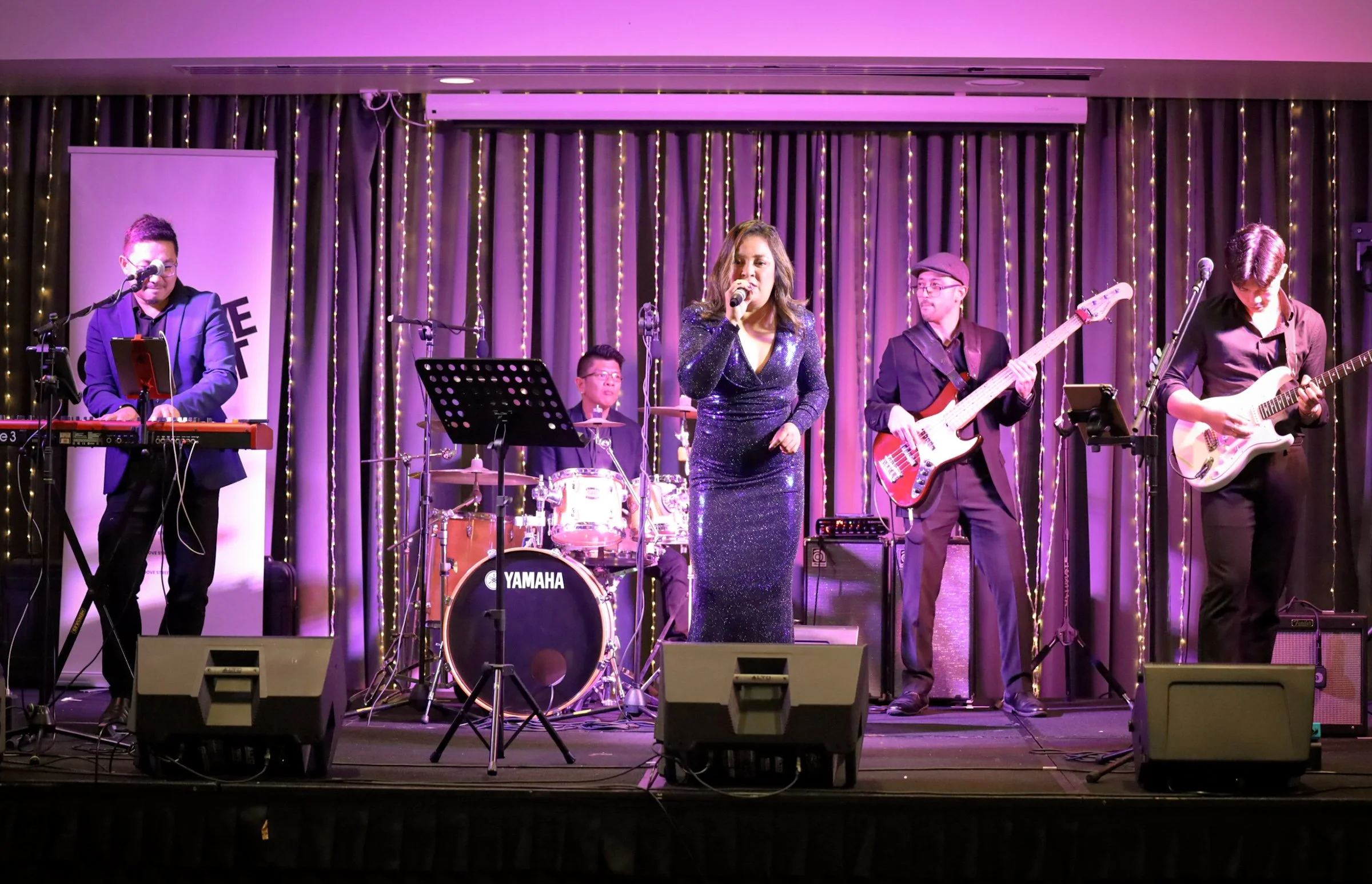 Band performing live on stage with purple and yellow lighting, including a female singer with a microphone, drummer, keyboardist, bassist, and guitarist.