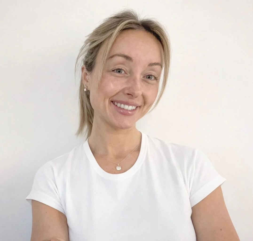Smiling woman with blonde hair in a white t-shirt standing in front of a plain white wall.