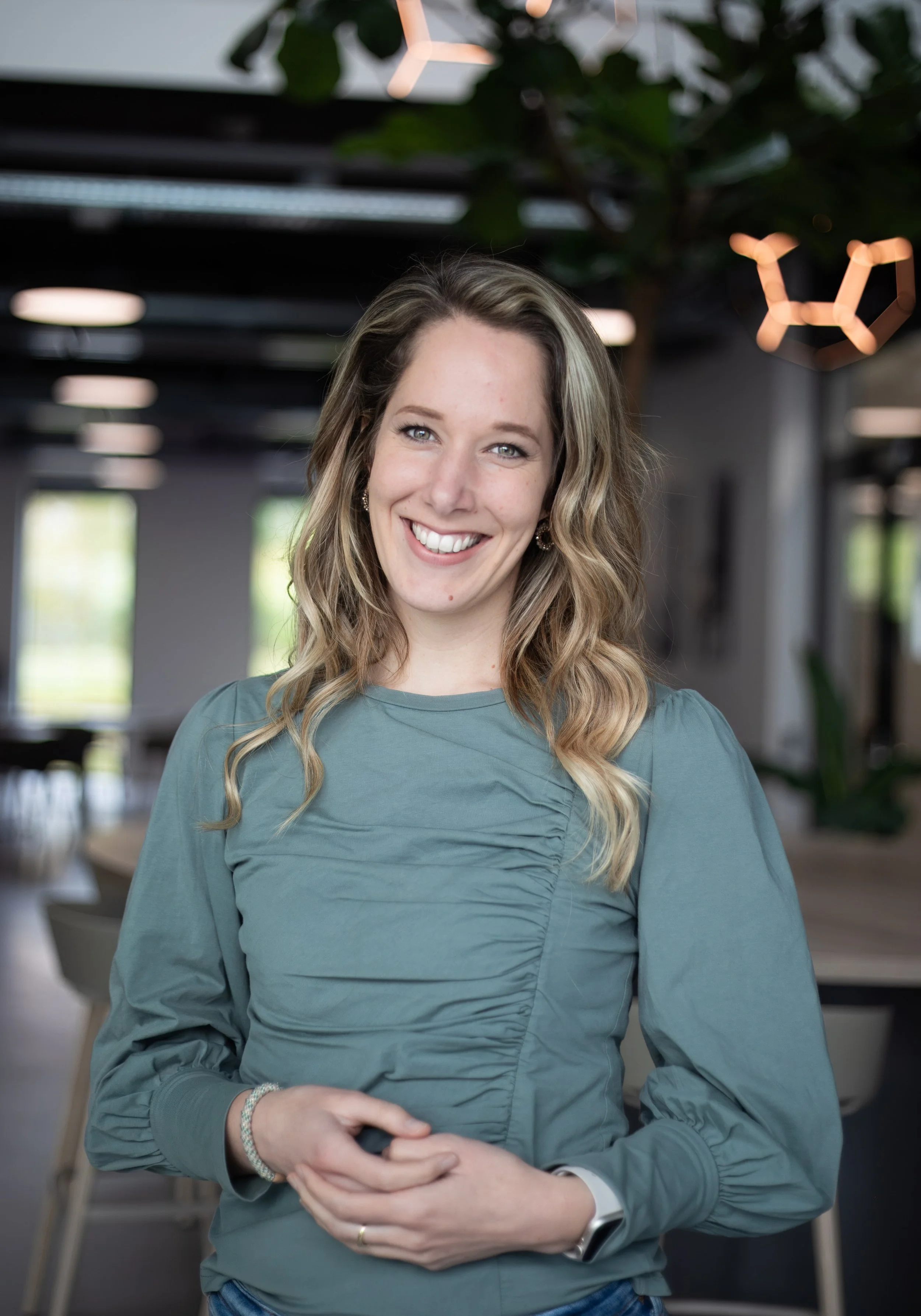 Lachende jonge vrouw met lange, golvende lichtbruine haren, knuffelend met haar handen, in een moderne kantoorruimte met groene planten en lichtdecoratie.