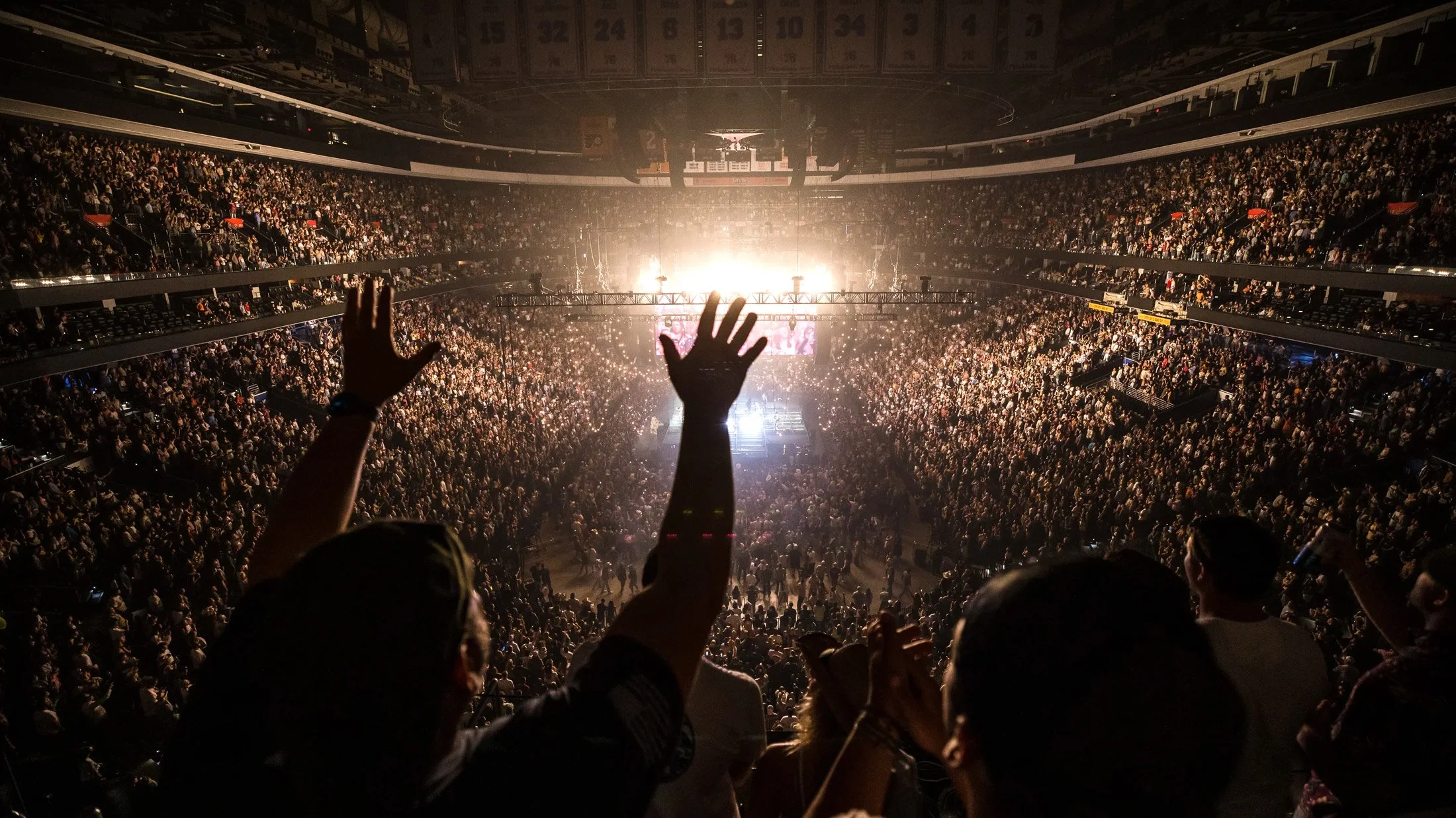 An indoor concert scene with a large crowd, stage lights, and audience members raising their hands.