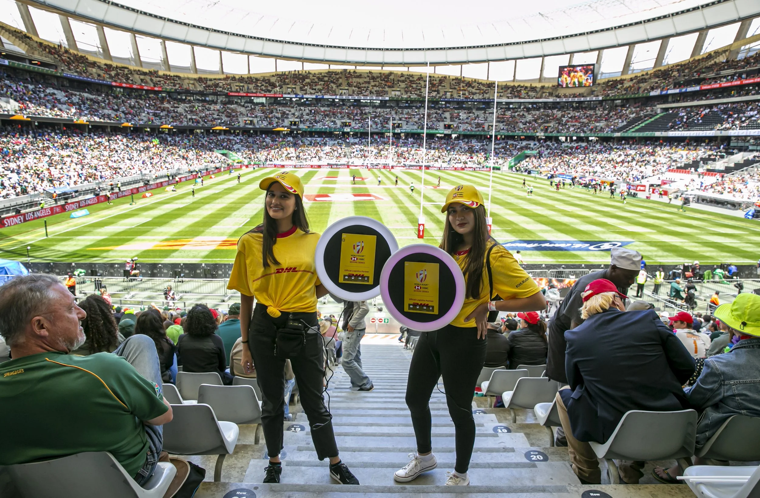 Two women wearing yellow shirts and yellow hats standing in a stadium, with a rugby field and spectators in the background.