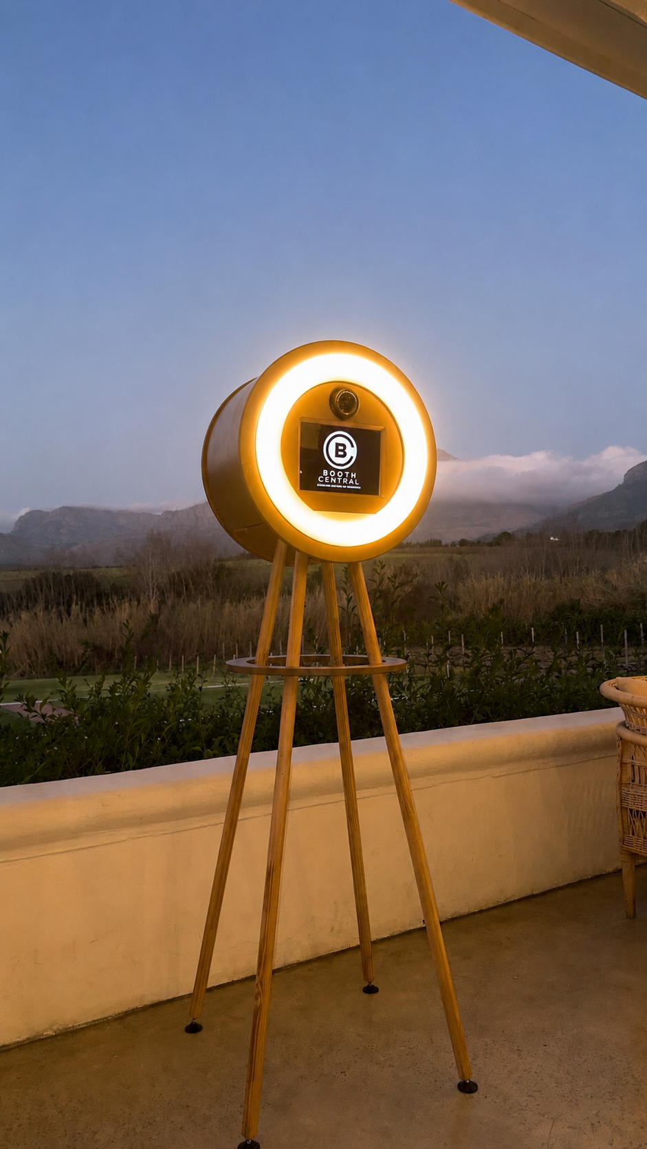 Photograph of a modern photo booth with circular LED lighting on a wooden tripod stand, displaying the logo for 'Booth Central', with a scenic outdoor background of mountains and trees during dusk.