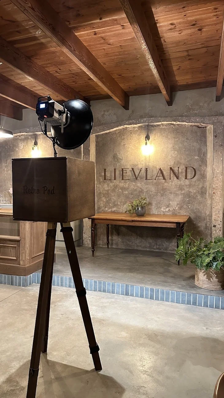 Interior of a rustic room with wooden ceiling beams, a textured concrete wall with the word "LIEVLAND" displayed, a wooden table with a potted plant, and a photo booth setup with a camera and a black reflector.