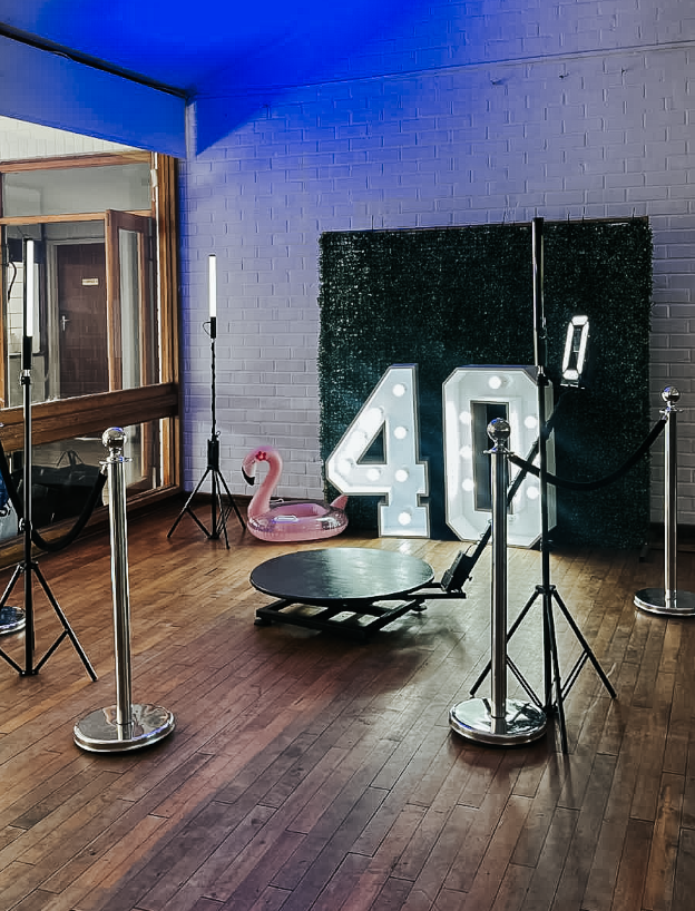 Indoor setup with large illuminated number 40, a pink flamingo float, and a black skateboard on a wooden floor, surrounded by stanchions with black ropes, near a windowed door with a blue ceiling and a brick wall background.