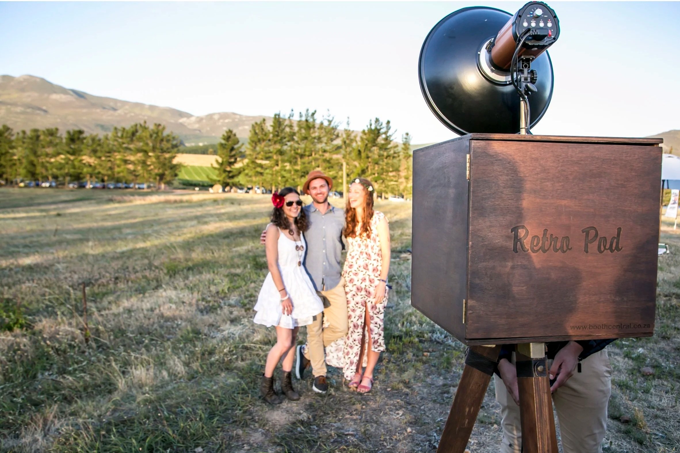 A group of three people, two women and one man, standing outdoors in a field with mountains and trees in the background, smiling and posing for a photo. In the foreground, a large retro camera on a tripod is visible.