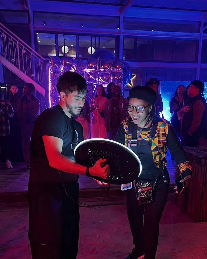 Two young adults looking at a tablet together in a lively, colorful party or event setting with other people in the background.