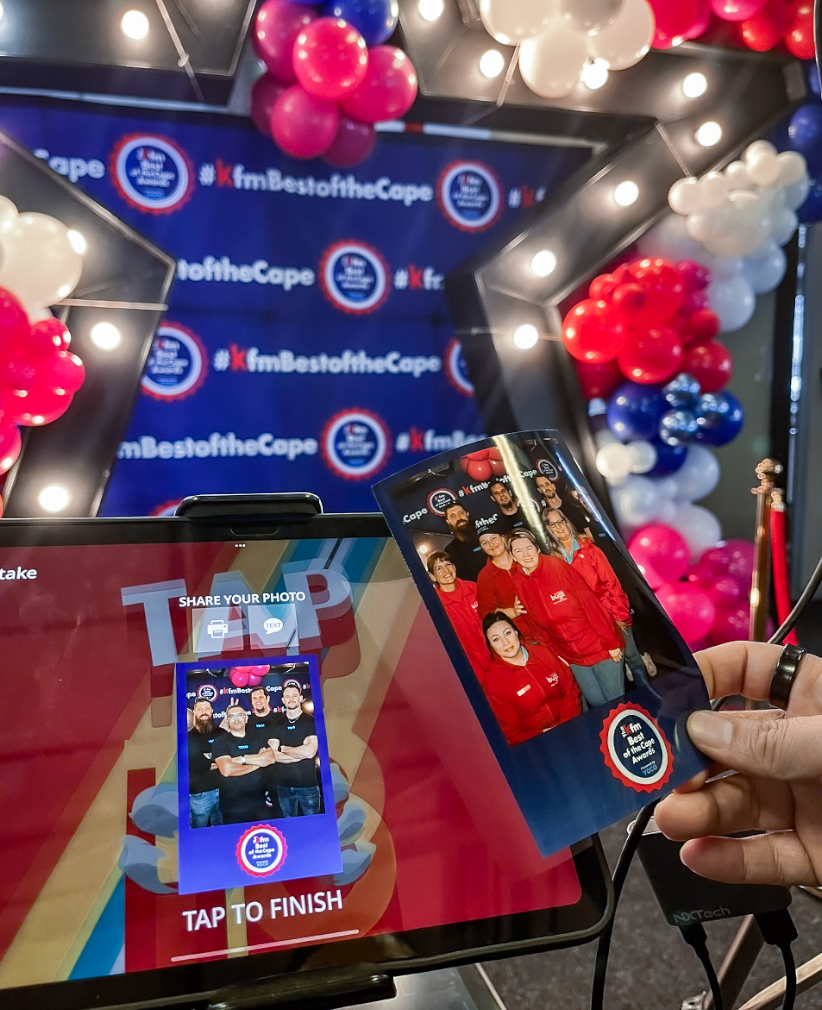 Photo booth setup with pink, red, white, and blue balloons framing an American-themed backdrop. A digital screen and a smartphone are showing group photos of people wearing red, with the backdrop displaying the hashtag #KfmBestoftheCape.