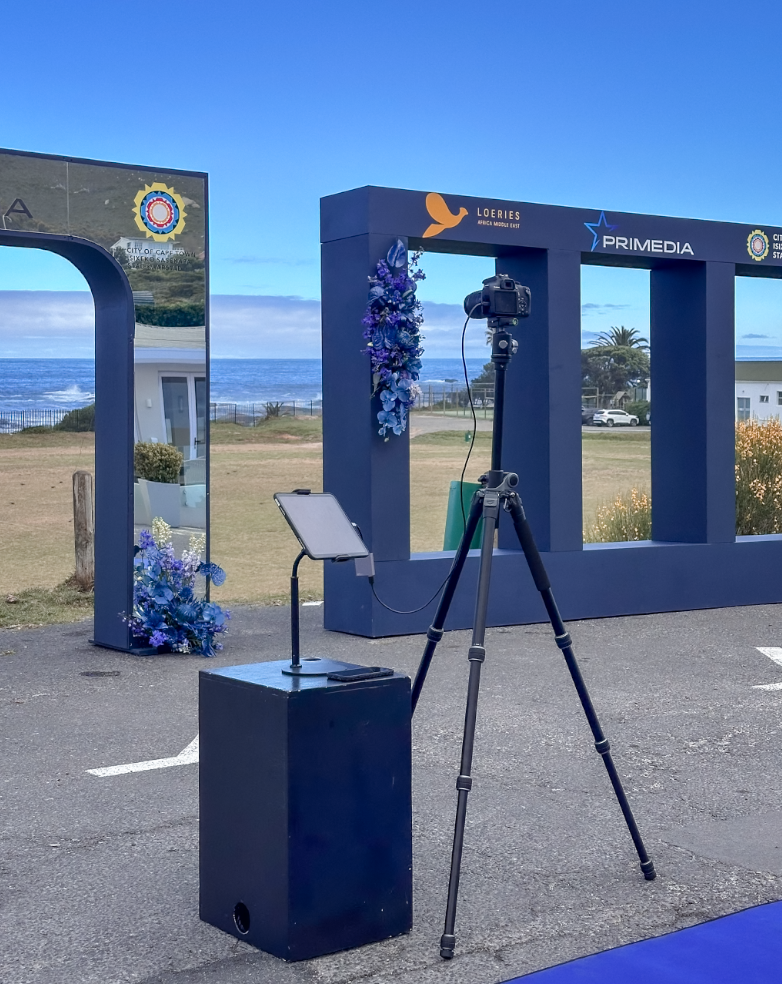 Camera on tripod capturing a scene with a decorative frame and floral arrangements, outdoor setting near the ocean with clear blue sky.