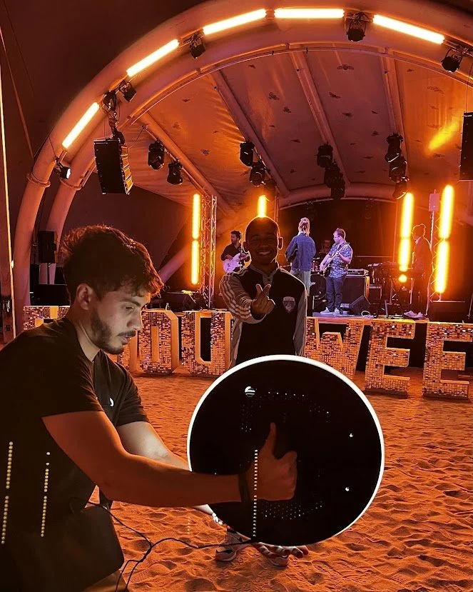 A young man is at a music event or concert, using a digital LED circle display to show visual effects. In the background, a band is performing on stage with orange vertical lights and musical instruments. The scene has a vibrant, energetic atmosphere with orange lighting.