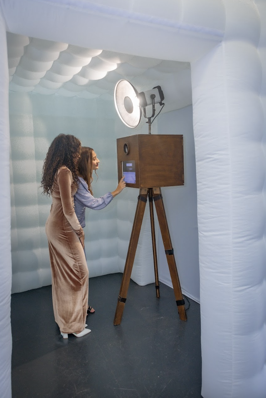 Two women are inside a white photo booth with a vintage wooden camera and a large flash on a tripod. One woman is operating the camera with a touchscreen, while the other looks on. The booth has soft, white, cloud-like ceiling padding.