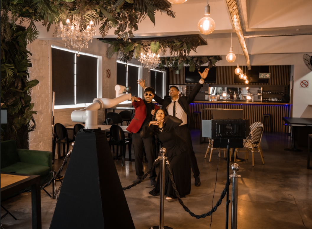 Three people dressed in formal attire taking a photo inside a decorated event space with greenery, chandeliers, and hanging lights.