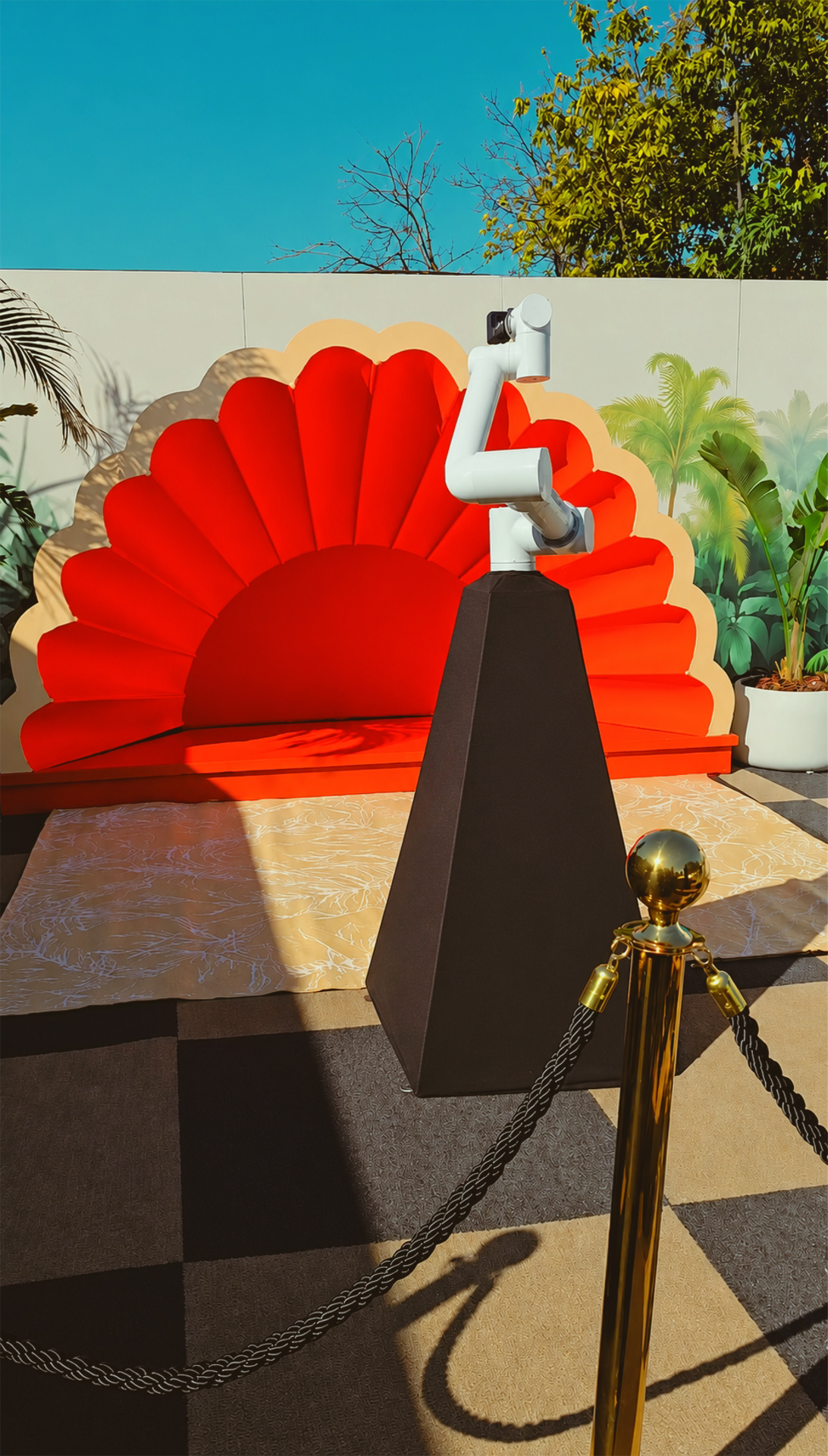 A red and cream fan-shaped decorative backdrop, a white robotic arm on a black stand, a bucket of lush green plants, a gold-colored stanchion with black rope, and a patterned mat on the ground under bright outdoor sunlight.