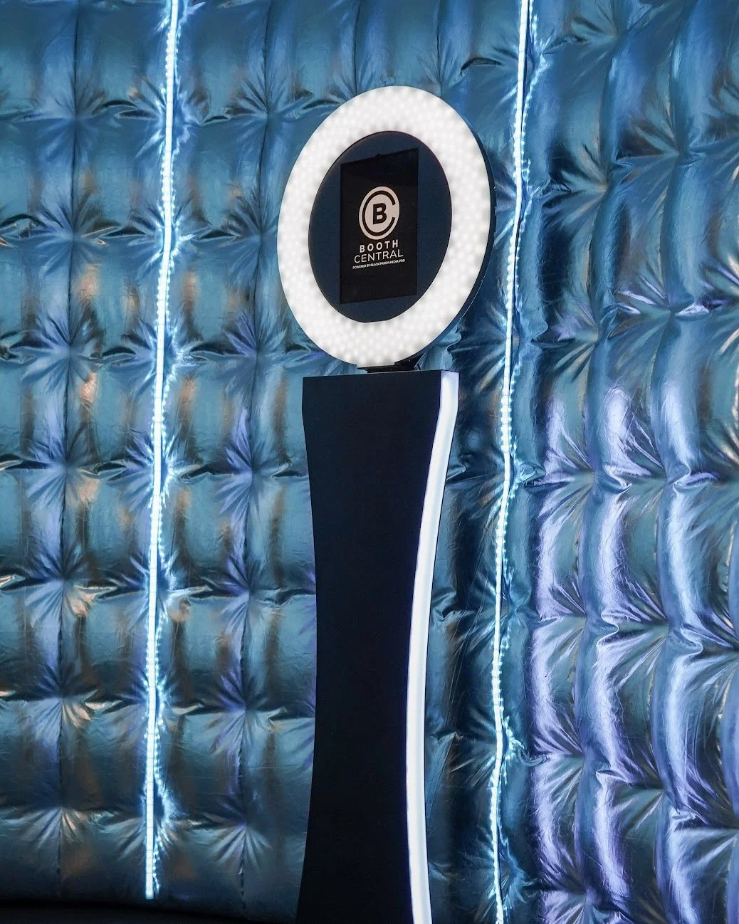 A digital photo booth with a futuristic design, featuring an illuminated oval mirror, a black pedestal, and a display screen reading 'Booth Central,' set against a shiny, quilted metallic blue wall with vertical blue LED strip lighting.