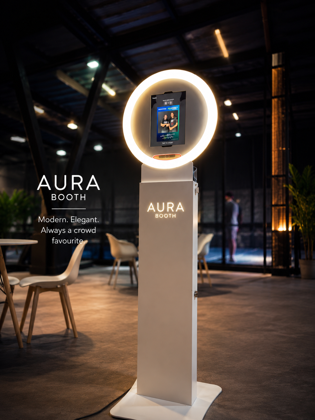 A modern photo booth named Aura Booth with a circular ring light and a digital screen for photos, set in a stylish indoor space with tables and chairs.