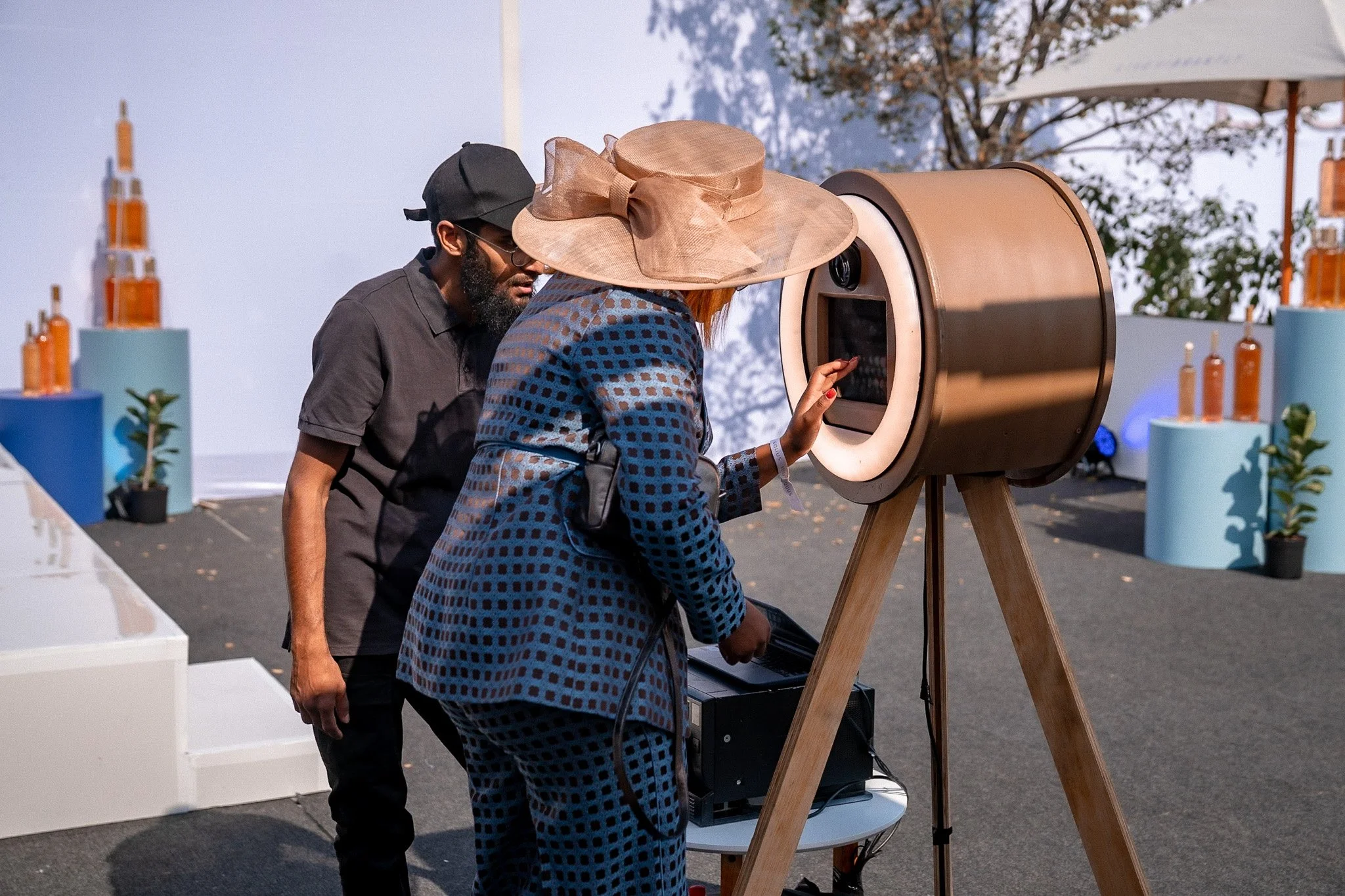 Three people, two women and a man, are looking into a large, horizontal, slit-like opening in a device that appears to be some sort of telescope or viewing instrument. The woman in the foreground is touching or adjusting the device. The other two are closely observing. They are outdoors, with trees and some structures in the background. One woman is wearing a large beige hat and a blue-patterned dress, the man is dressed in black and wearing glasses, and the other woman is wearing a black cap and dark clothing.