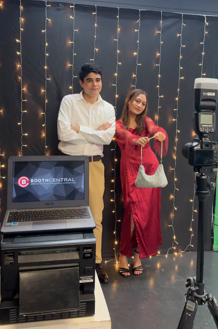 Two people, a young man and a woman, stand in front of a black backdrop illuminated with string lights. The woman is holding a silver purse and appears to be posing for a photo. There is a camera on a tripod and a computer screen displaying 'Booth Central' in front of them.