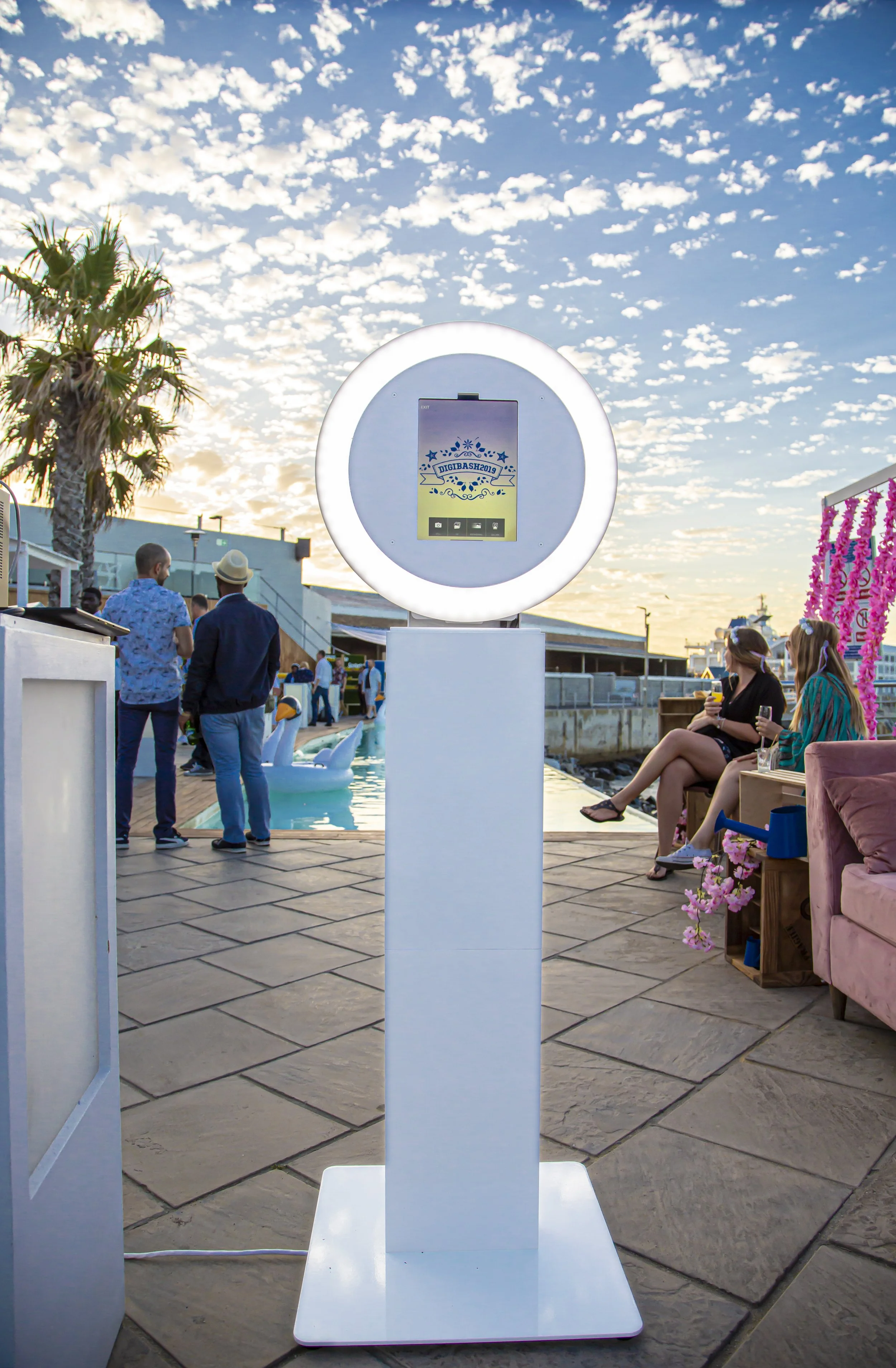 A white, illuminated round ring light with a rectangular stand in an outdoor setting during sunset. In the background, there are people gathered, some standing and some sitting, near a pool with swan-shaped inflatables. The area is decorated with pink flowers and pink streamers, with a palm tree on the left and a clear sky with scattered clouds.