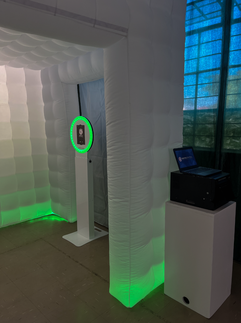 A booth with a white, inflatable, padded structure with green lighting at the base, a digital screen on a stand with a contactless payment icon, and a laptop on a white pedestal, situated by a window with blue blinds.