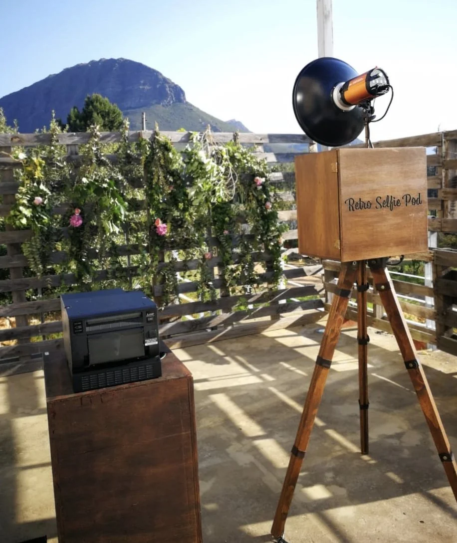 A retro selfie photo booth setup on a balcony with a wooden box labeled 'Retro Selfie Pod,' a camera inside a large black camera housing, and a small printer on a wooden stand, overlooking mountains and a garden with pink flowers.
