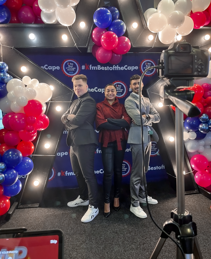 Three people standing in front of a backdrop with #KfmBestoftheCape, surrounded by colorful balloons, at a photo booth for a radio awards event, with a camera set up to take their picture.