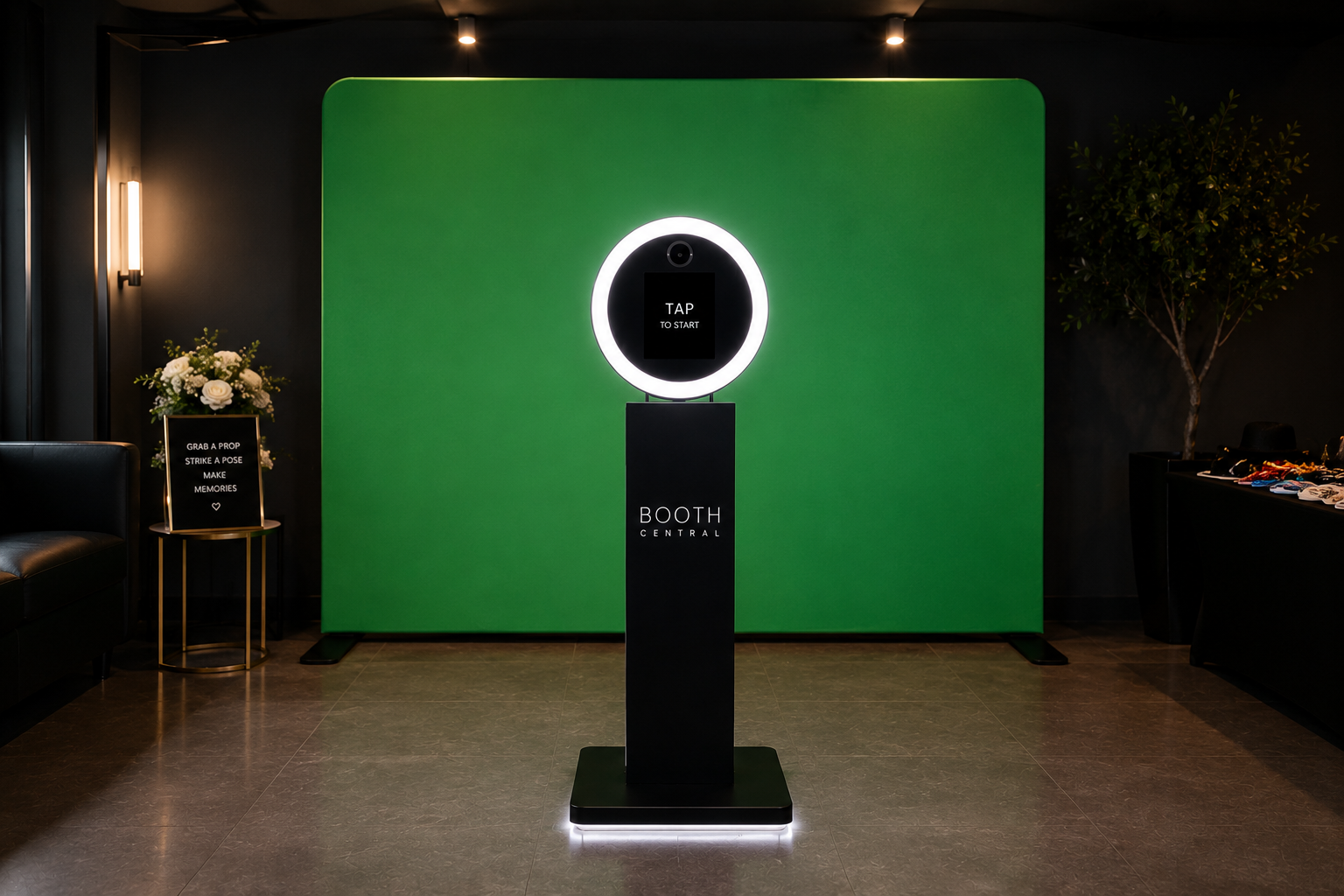 A photo of a modern photo booth setup with a black stand, a circular ring light, and a screen that says "TAP TO START." The background is a large green wall, and to the left, there's a small table with a floral arrangement and a sign that reads "Grab a prop, strike a pose, make memories." To the right, there is a black table with various props and a potted tree.