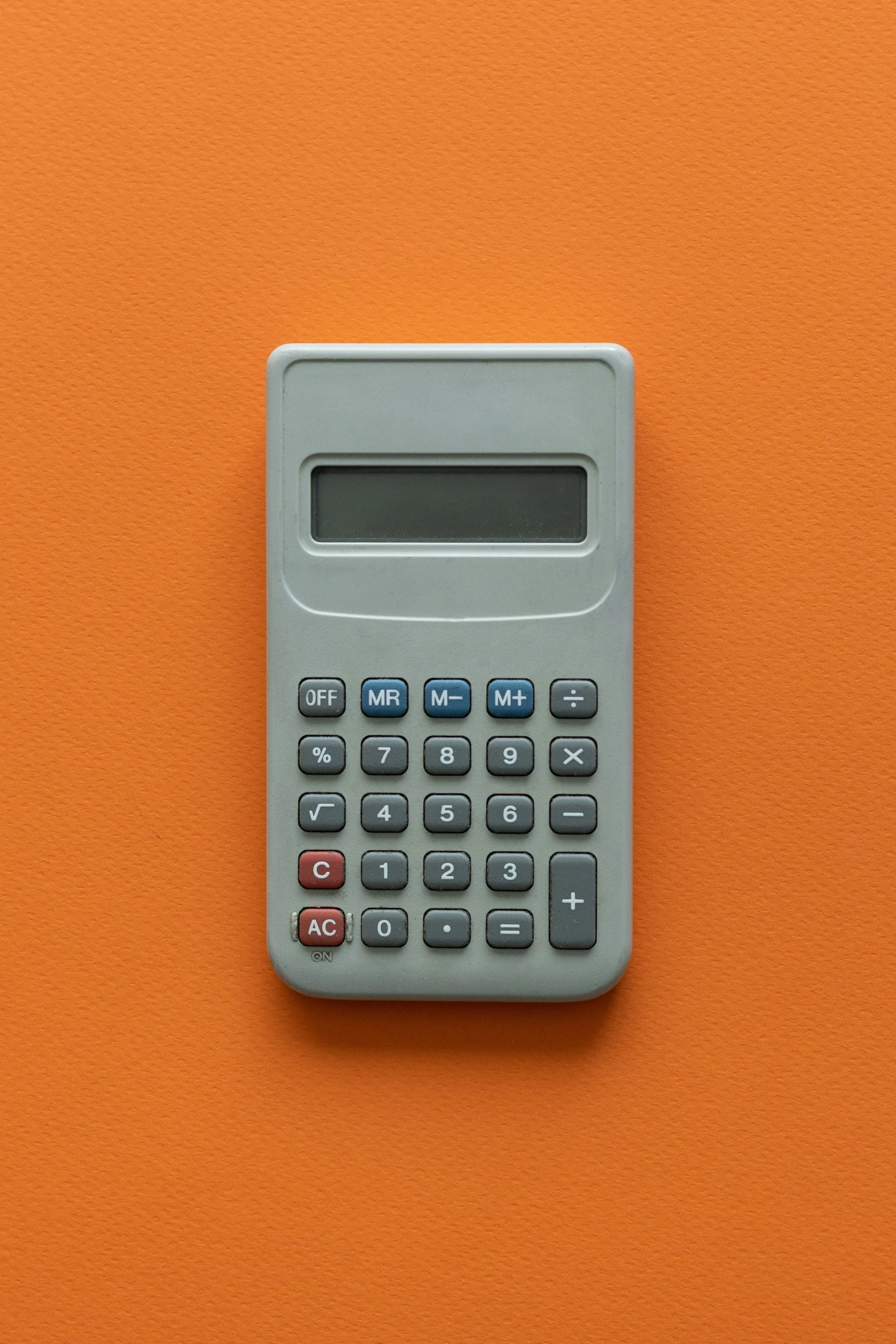 A gray calculator with a black display and various gray and red buttons, placed on an orange surface.