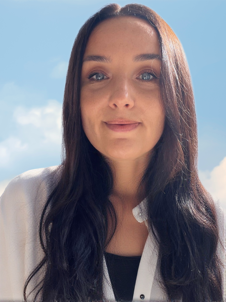 Photo de Emmanuelle Laisney, la graphiste. Elle a les cheveux longs et bruns sourit légèrement, portant une chemise blanche sous un ciel bleu avec quelques nuages.
