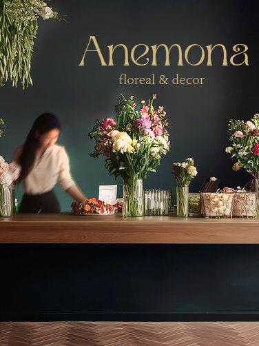 Une réception avec plusieurs arrangements floraux colorés sur un comptoir en bois, avec une personne en arrière-plan manipulant des fleurs, et un panneau indiquant 'Anemona floral & decor'.