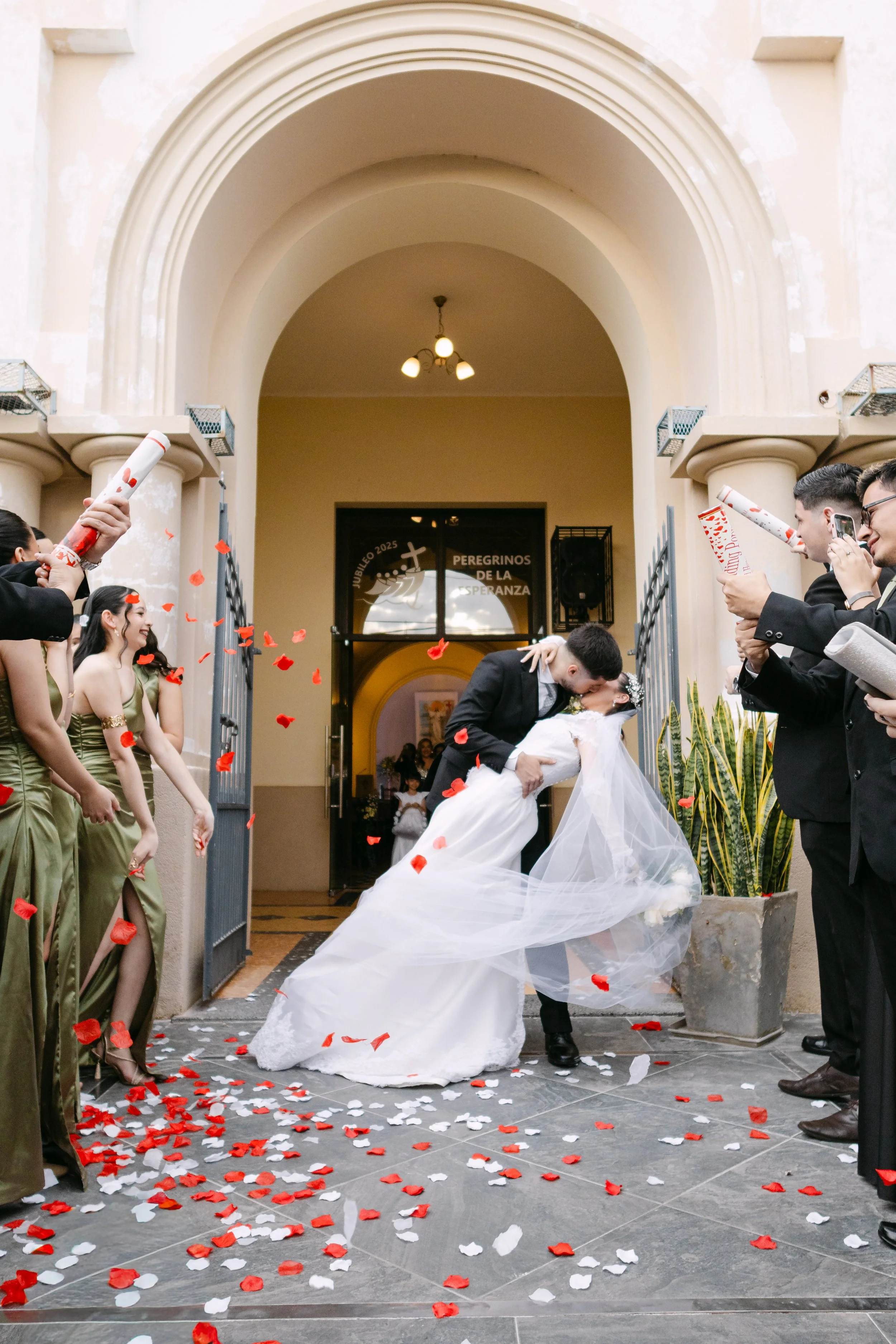 Una pareja de novios besándose en la entrada de una iglesia durante su boda, rodeados de amigos y familiares que lanzan pétalos de rosa blancos y rojos.