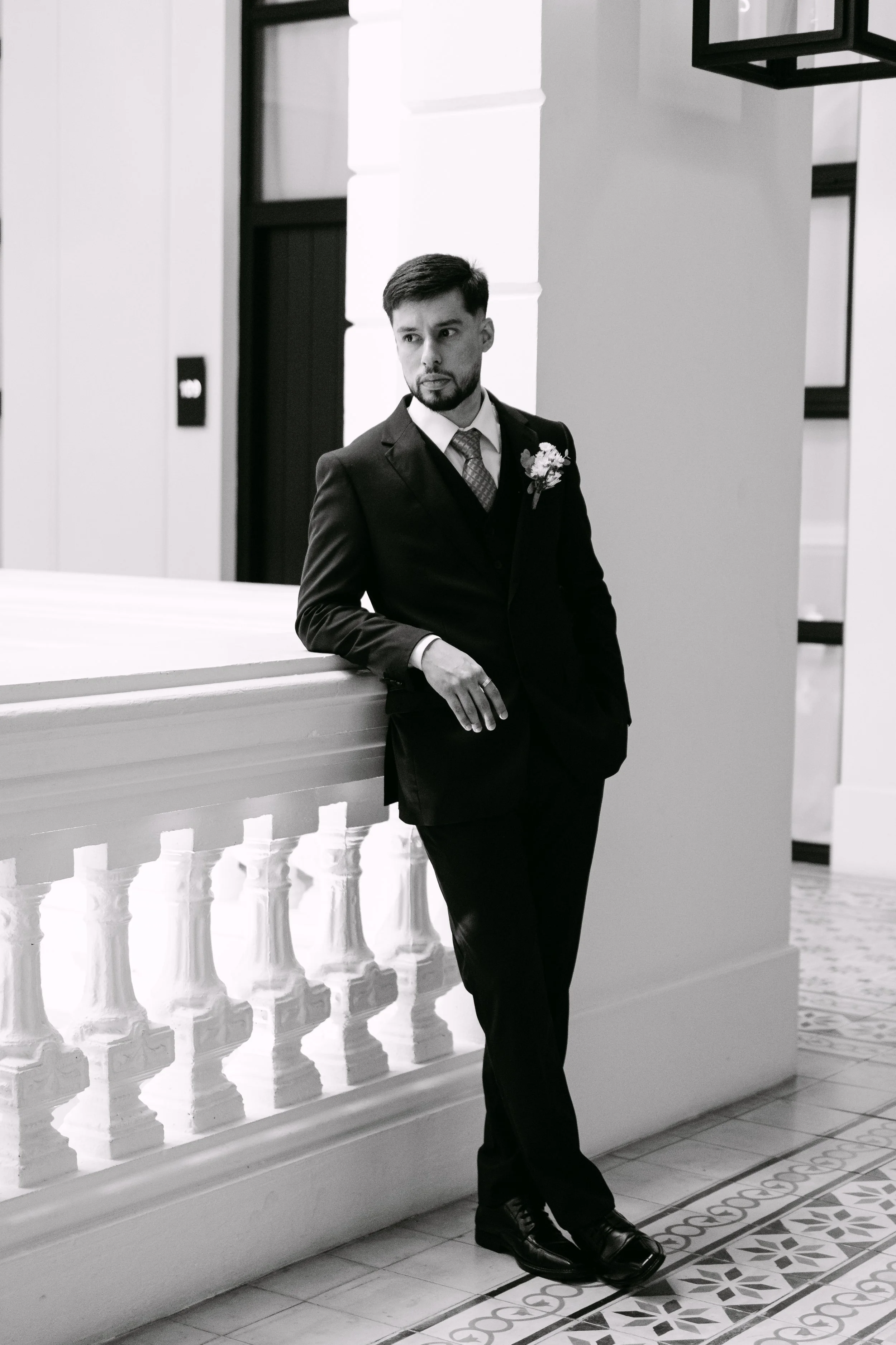 Hombre de negocios con traje negro y corbata, de cabello castaño oscuro, con barba, apoyado en una barandilla blanca en un interior elegante con paredes blancas y decorativas, y suelo de azulejos con patrones.