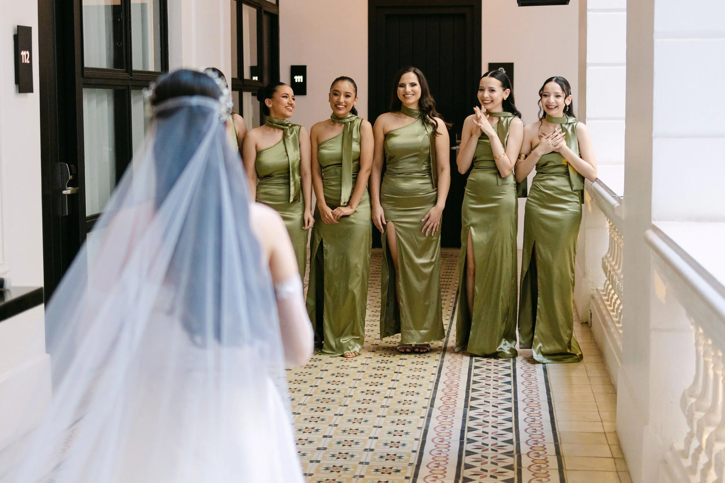 Un grupo de seis mujeres vestidas con trajes largos y elegantes, de color verde, están en un pasillo y parecen estar esperando para un evento especial, mientras una mujer con vestido de novia en el primer plano las observa.