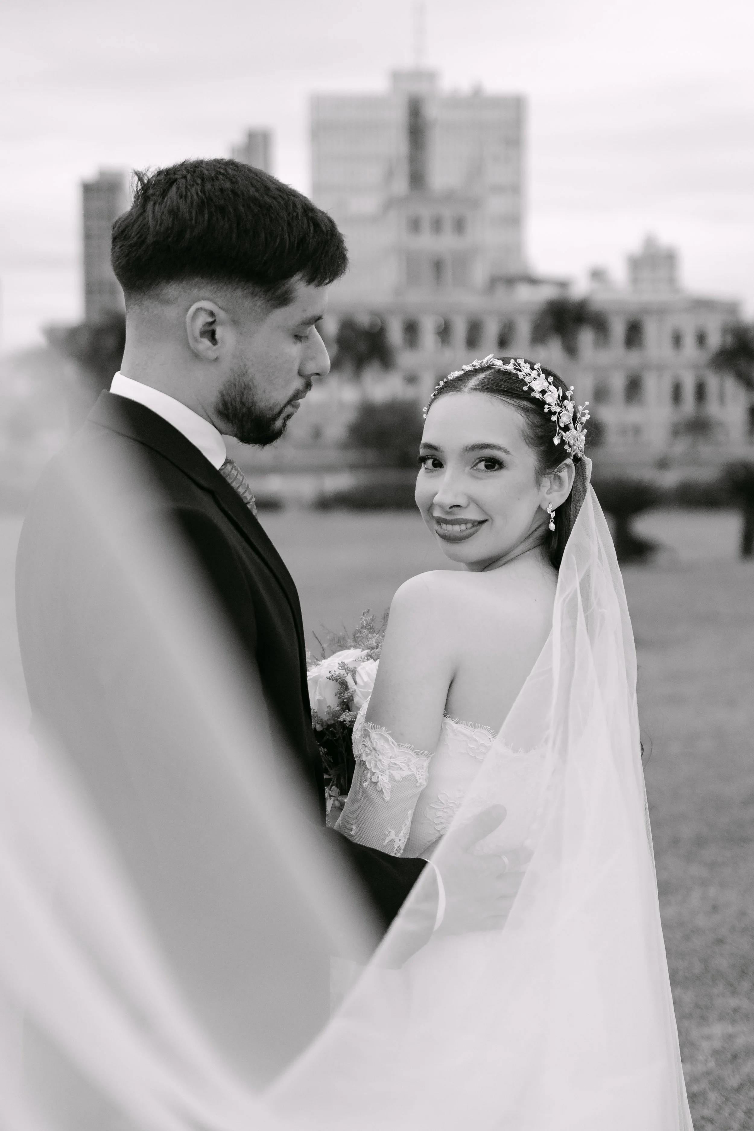Una pareja de novios en una boda, en blanco y negro, con la novia sonriendo y luciendo un vestido de boda y una corona de flores en el cabello, mientras el novio viste un saco y una corbata. Están en un espacio exterior con edificios de la ciudad en 