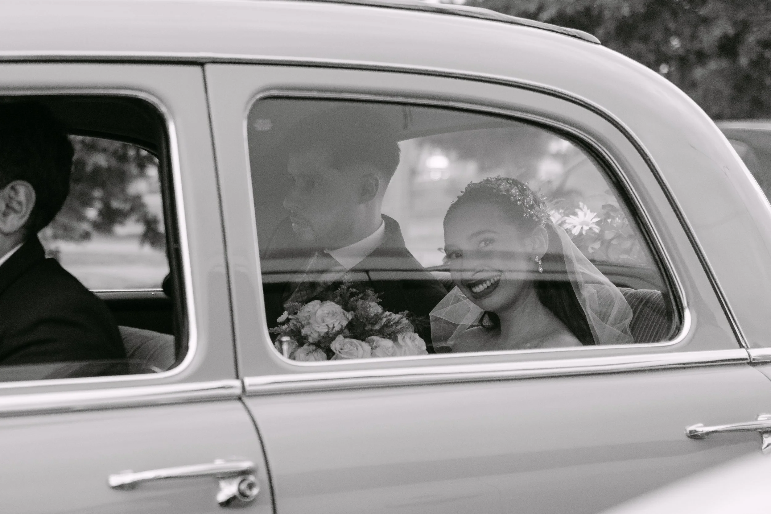Una pareja boda viaja en un coche clásico, ella lleva un vestido de novia y sostiene un ramo de flores, ambos sonríen y parecen felices.