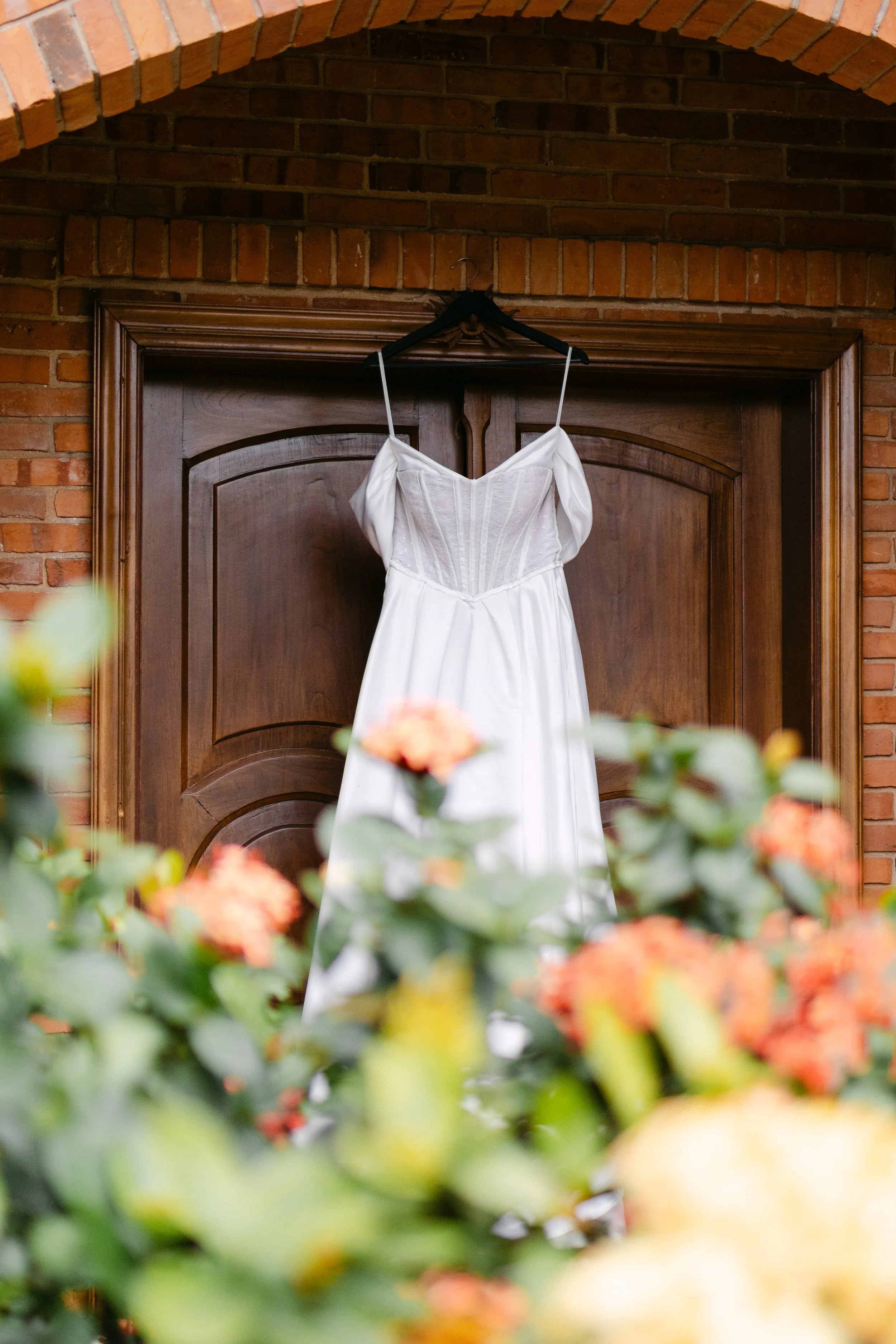Vestido de novia blanco colgado en una puerta de madera, con fondo de pared de ladrillos y flores en primer plano.